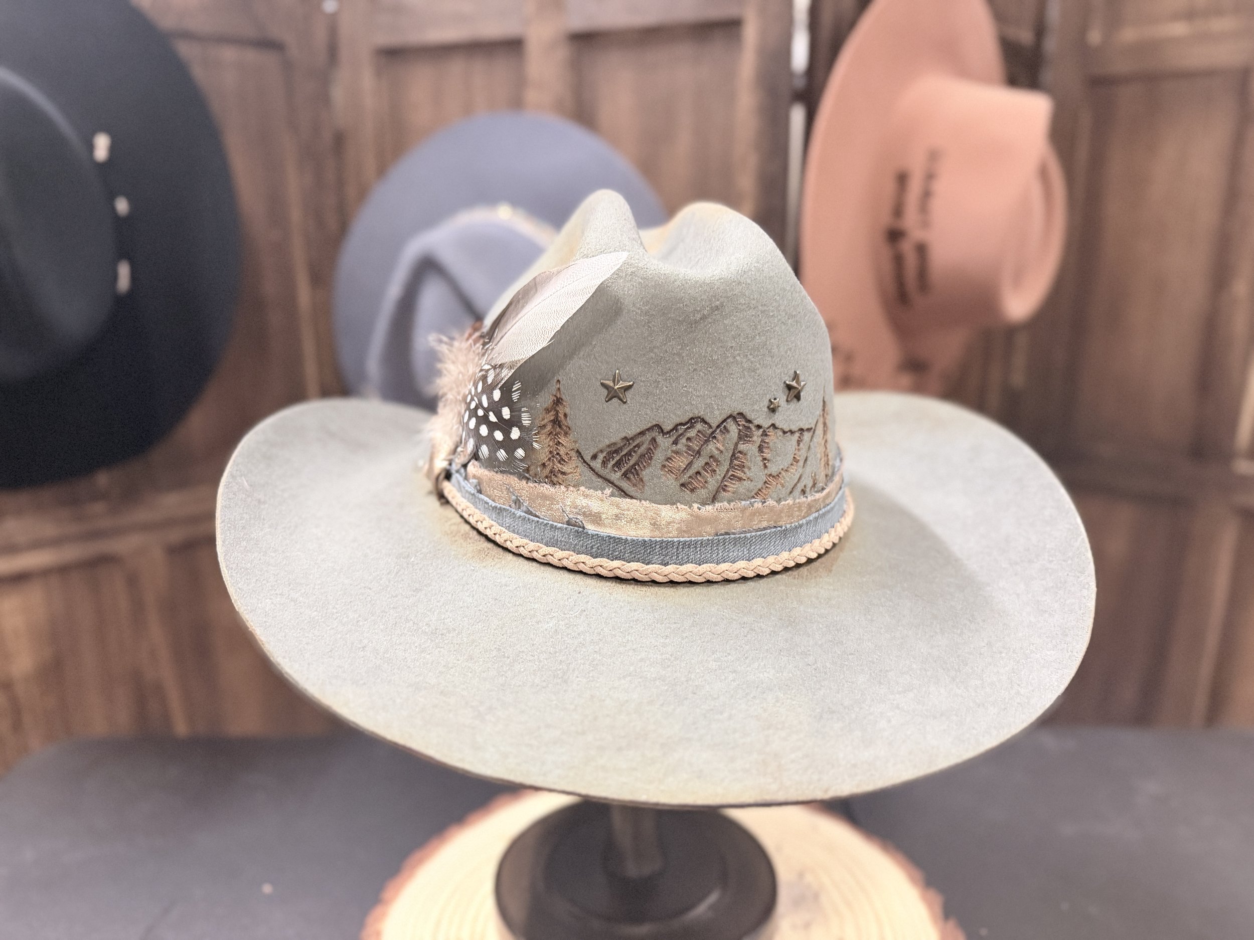 The image shows a decorative beige hat with mountain and star designs, placed on a beige surface. In the background, there are two other hats, one black and one peach, on a wooden surface.