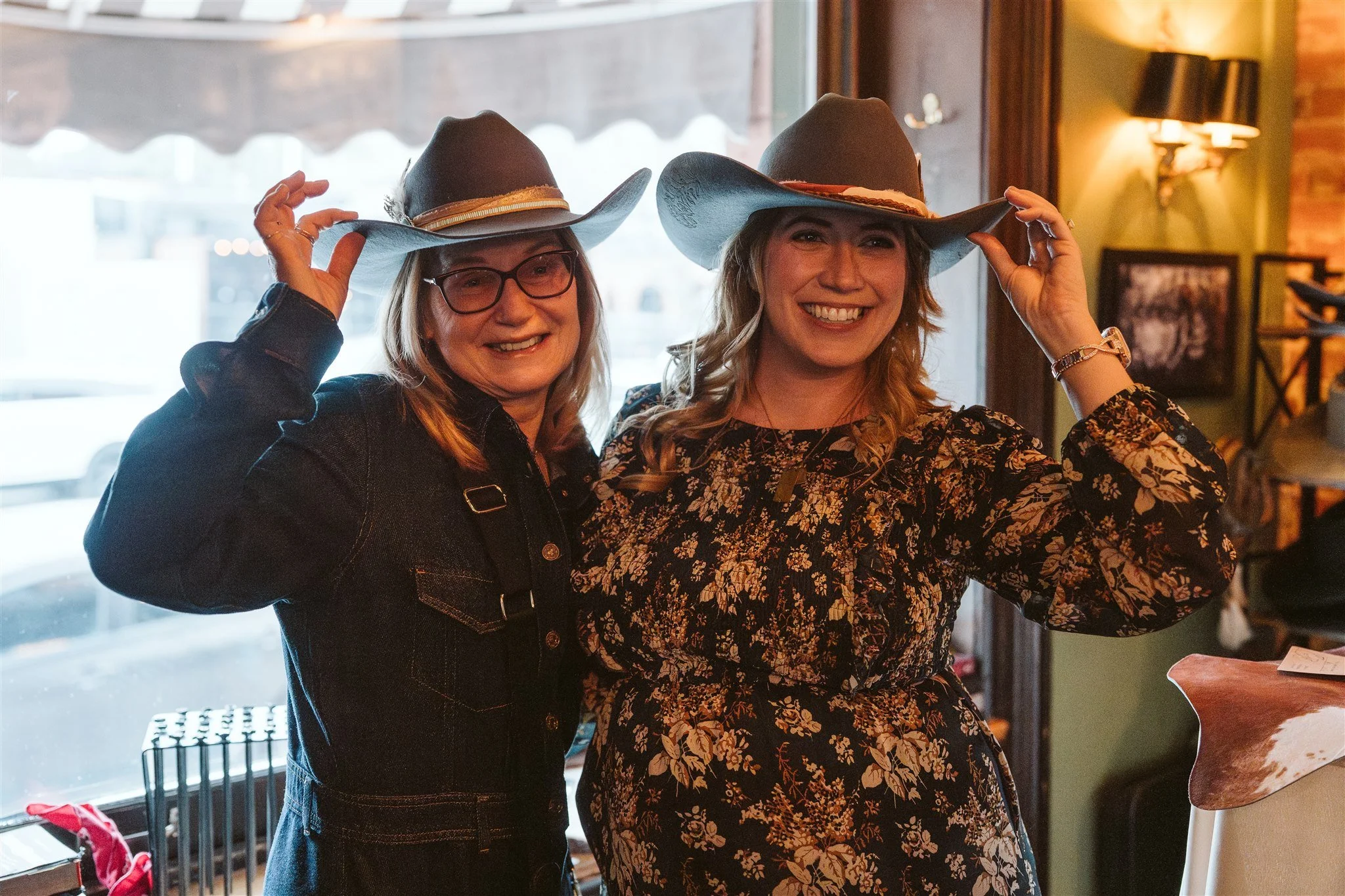 Two women wearing cowboy hats and smiling inside a warmly lit room with a window and green walls.