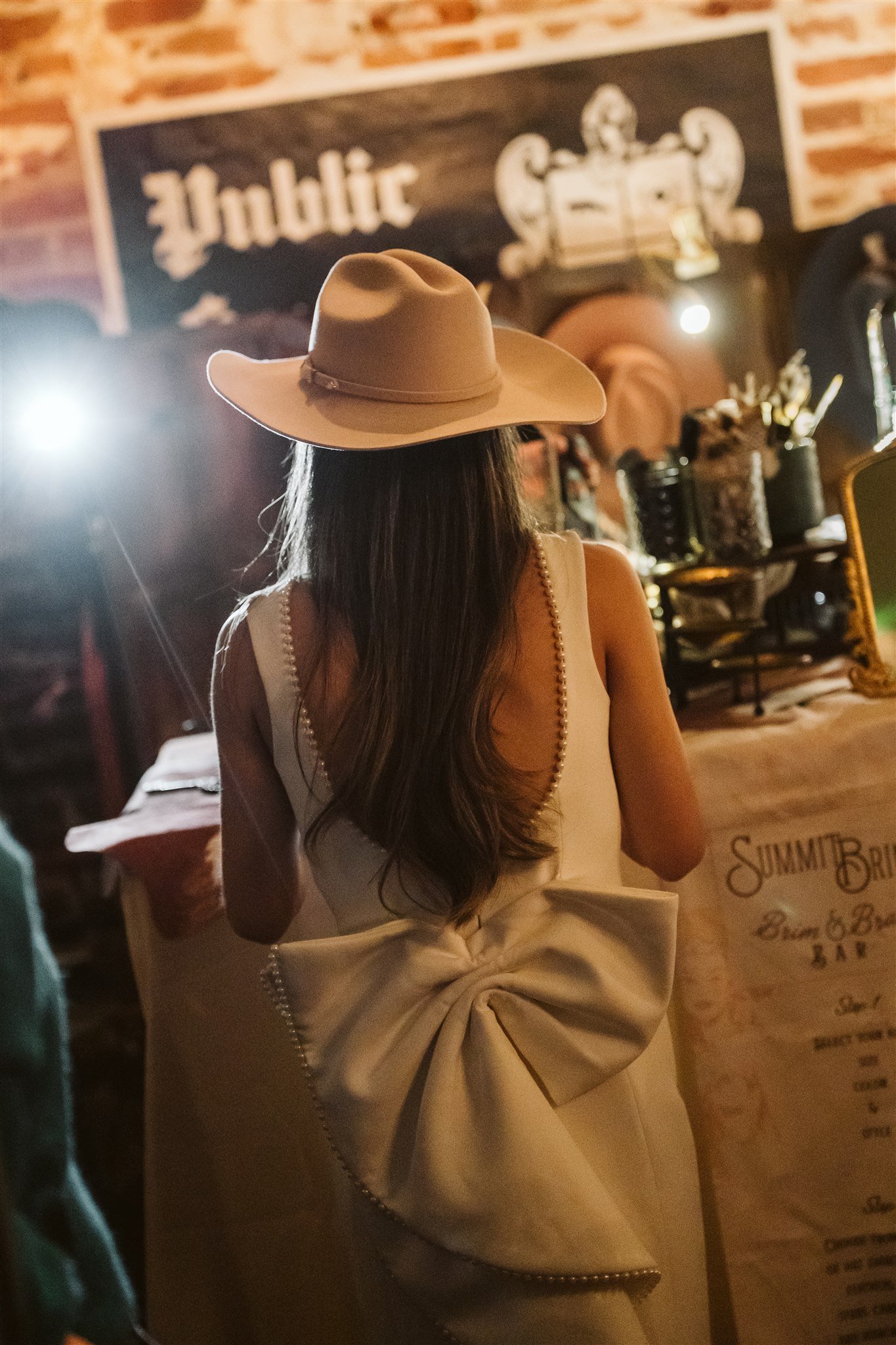 A woman with long hair wearing a beige wide-brimmed hat and a sleeveless dress with pearl detailing, standing in a rustic bar or restaurant with exposed brick walls, a chalkboard sign, and wooden furniture.
