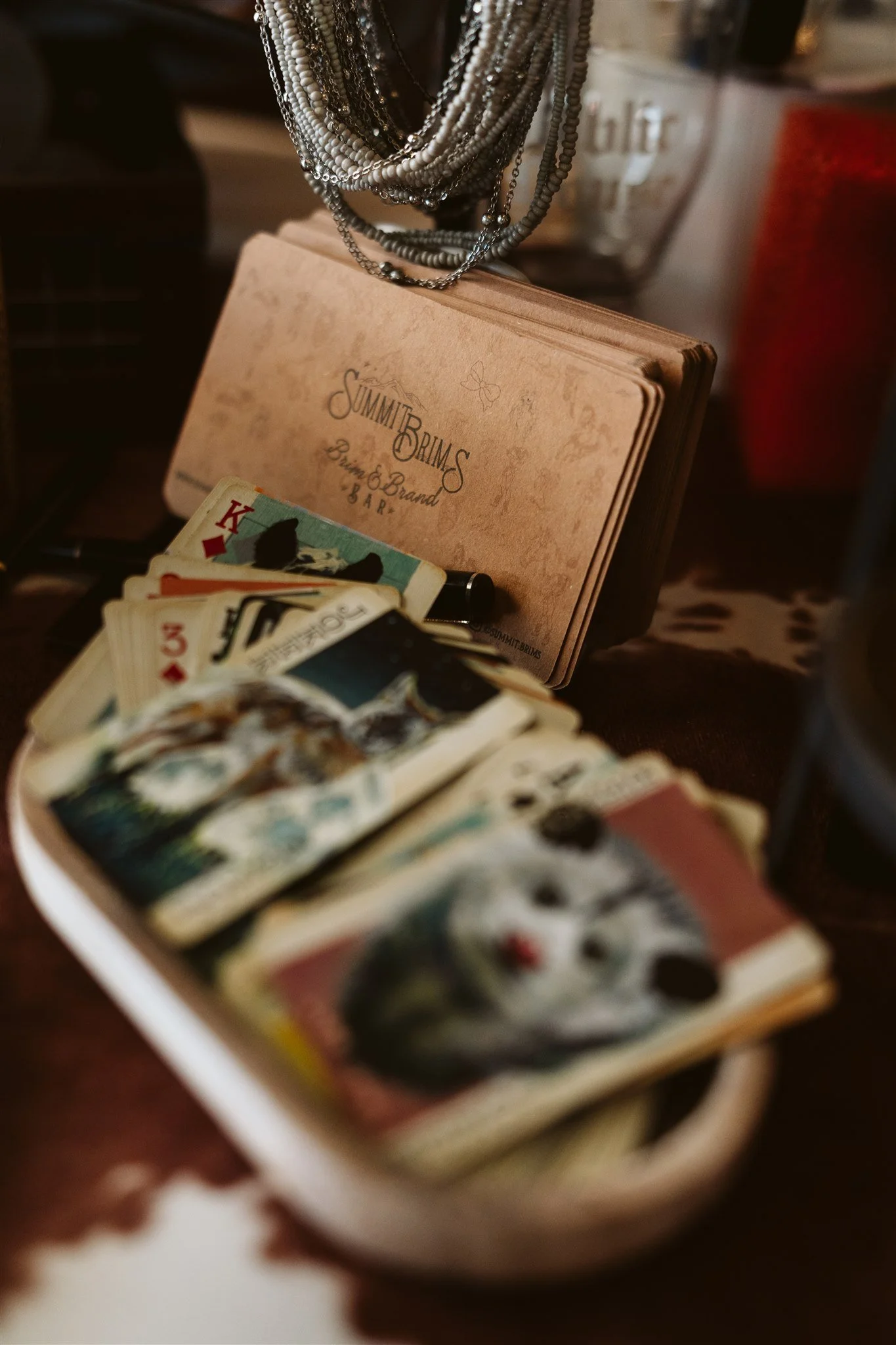 Close-up of a cup of playing cards, some face cards visible, with a bowl of money in the background, a book titled 'Summit Brims' with a necklace draped over it behind the cards, and a glass container with the word 'love' written on it to the right.