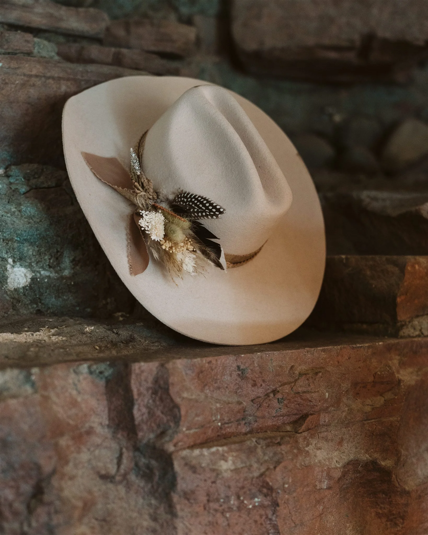 A white cowboy hat with feather decoration resting on a rough brick wall.
