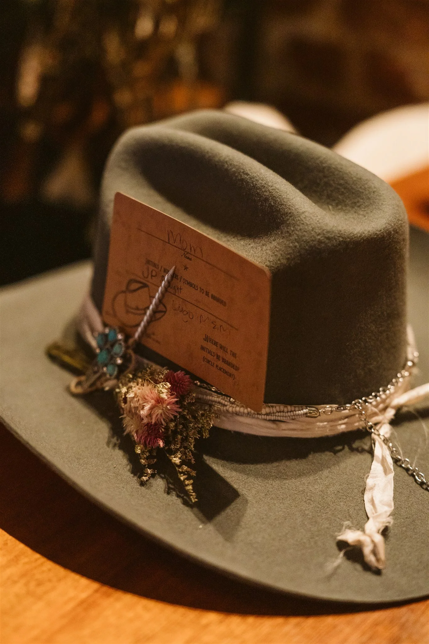 Close-up of a small black hat with a pink jewelry tag attached, surrounded by jewelry and dried flowers, on a black surface.