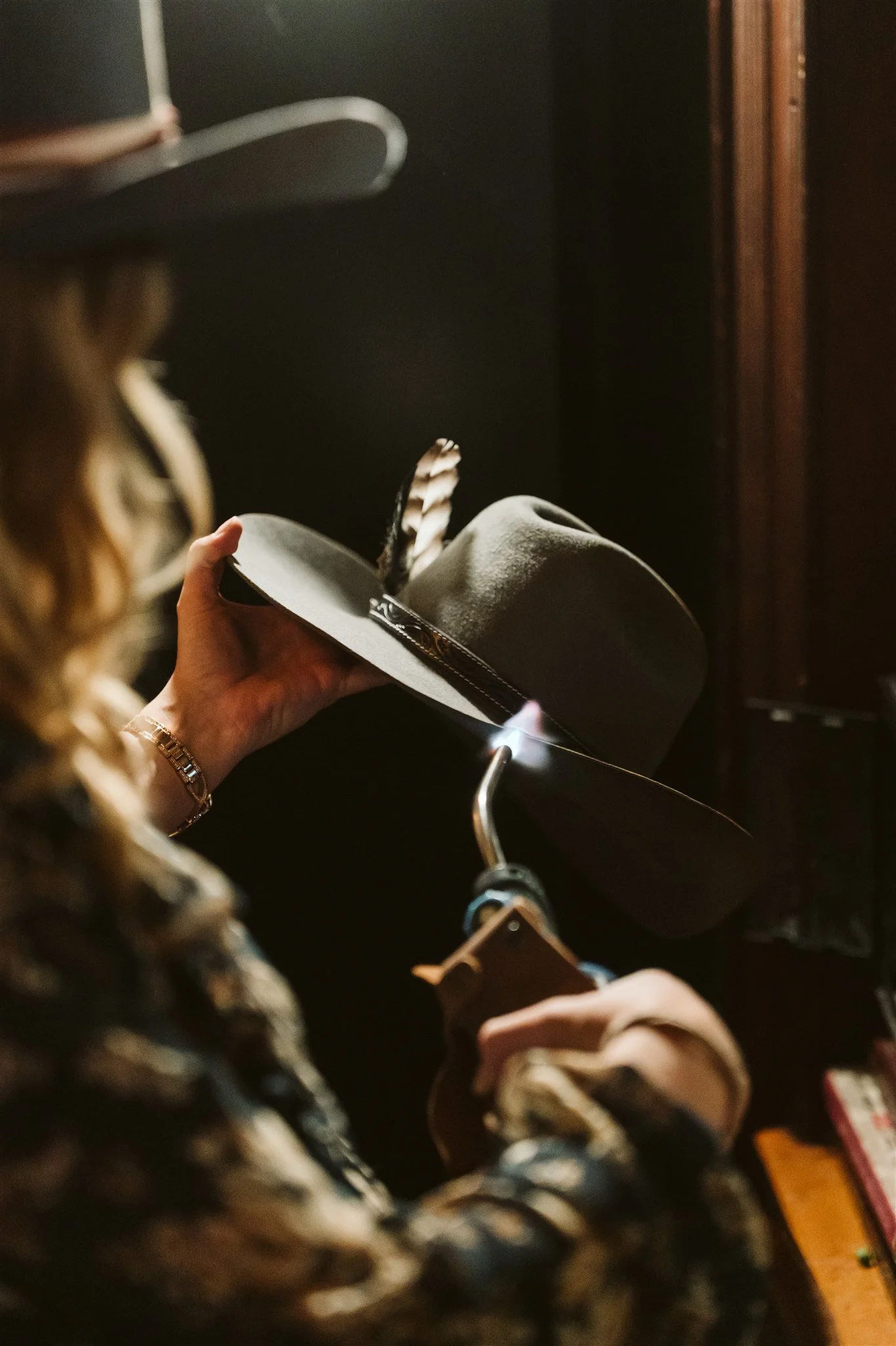 A person holding a gray fedora hat with a feather detail inside. The background shows a dark interior space with wooden elements and a metal hook.