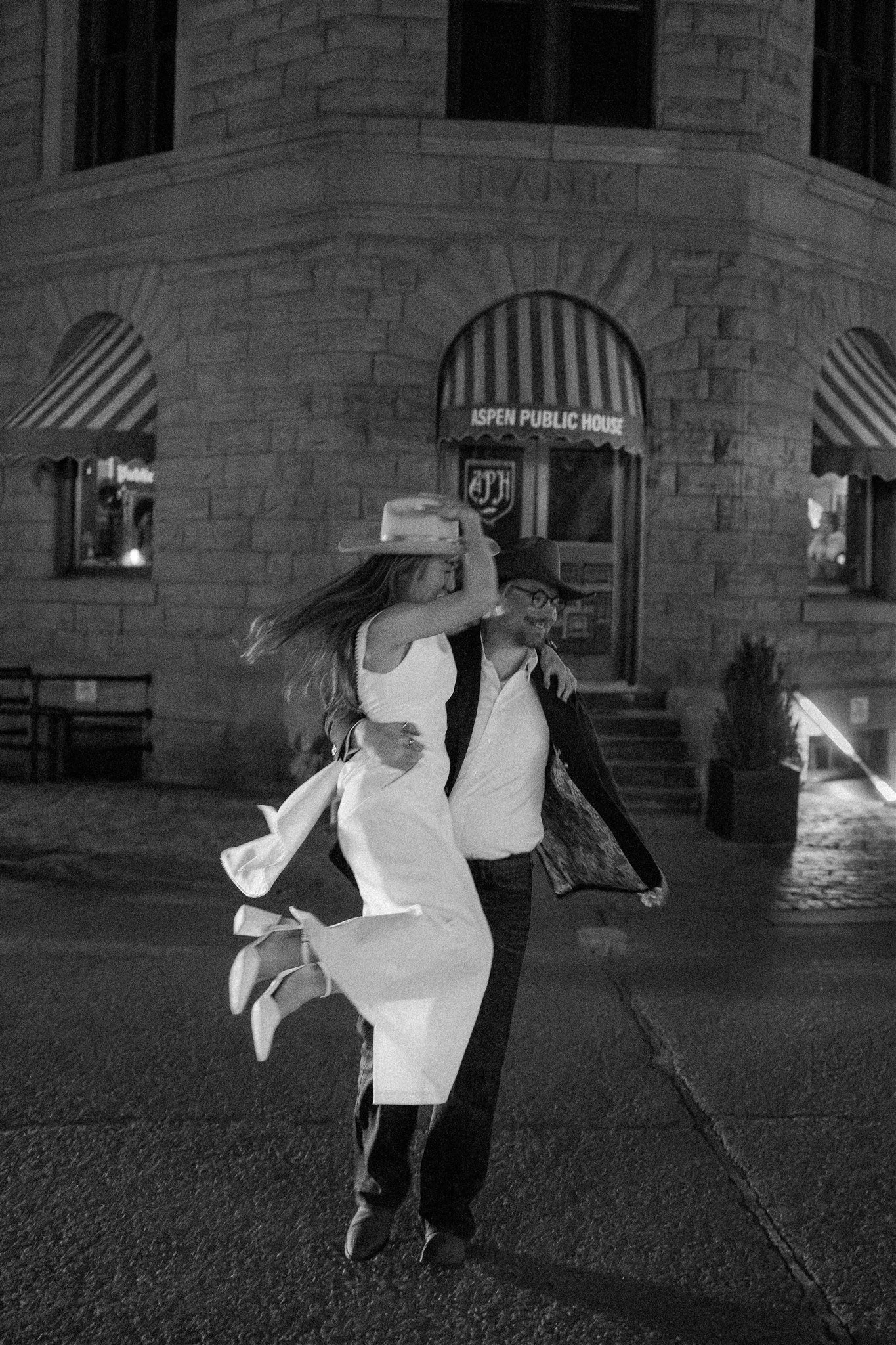 A man and a woman are dancing outdoors at night, the woman appears to be in a white dress with a wide-brimmed hat, and the man is wearing a hat and dark clothing. They seem to be enjoying a lively moment.