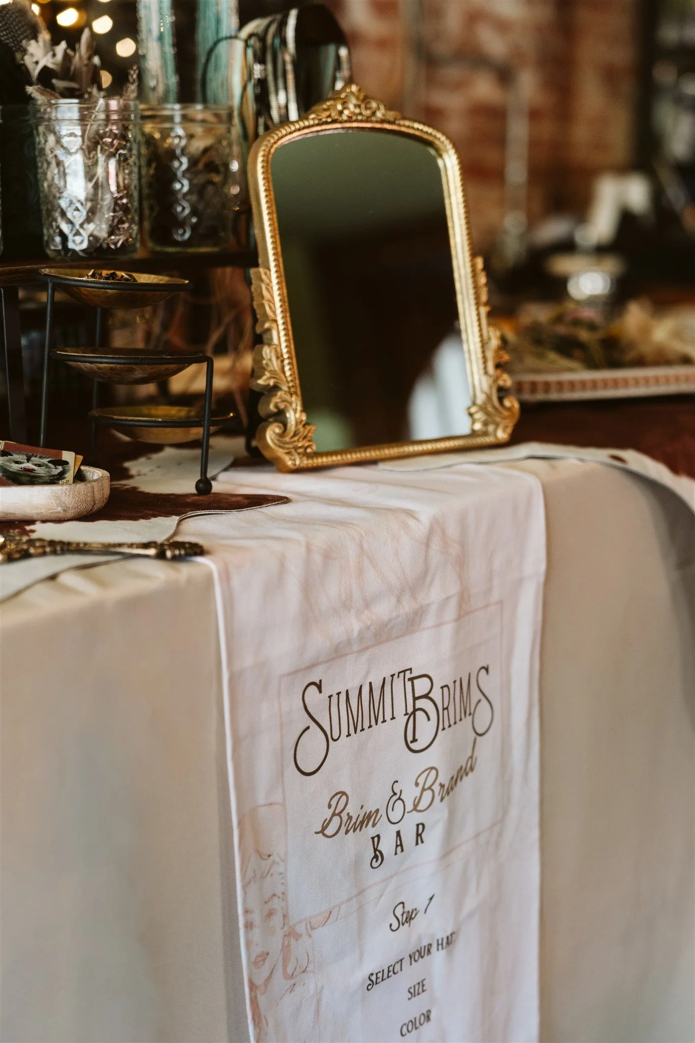 Decorative table display with a gold-framed mirror, glass jars filled with small items, and a white cloth banner with the text 'Summit Brims' and other text partially visible, indicating a branding or product display setup.