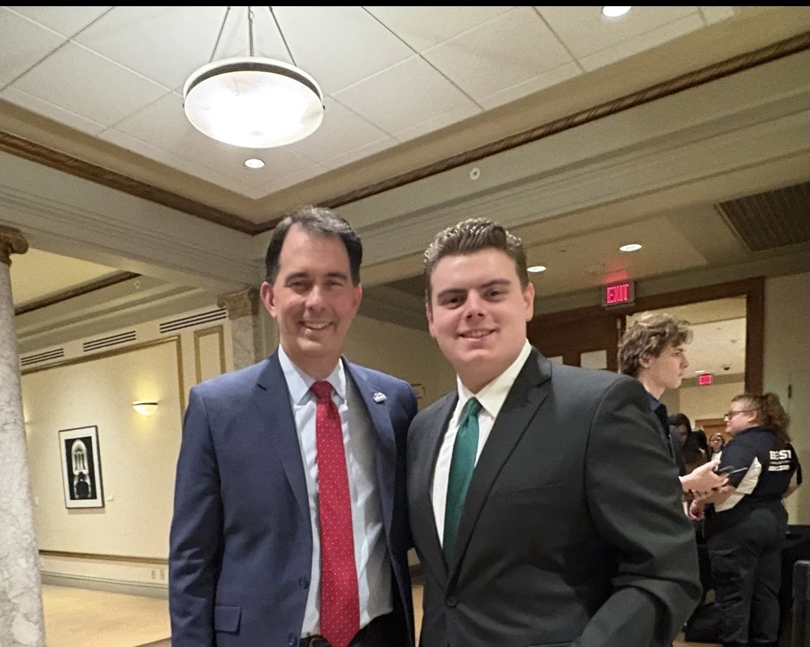 Two young men in suits standing together at an indoor event, smiling at the camera.