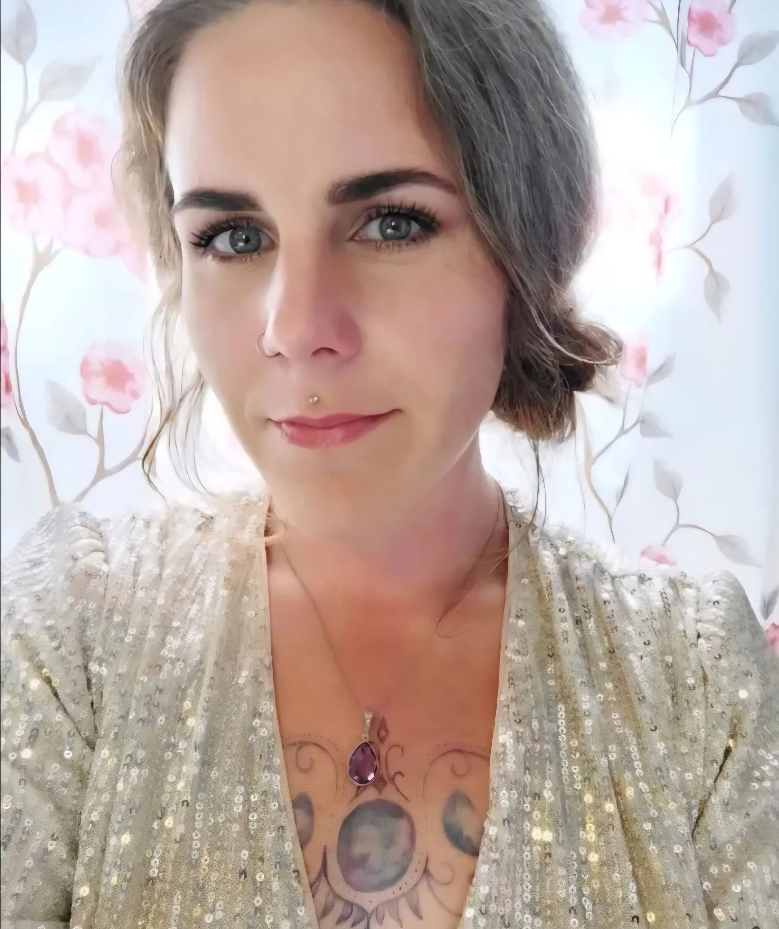 Close-up of a woman with gray and brown hair, wearing a sparkly silver top, a purple pendant necklace, and tattoos on her chest, standing in front of a floral wallpaper background.