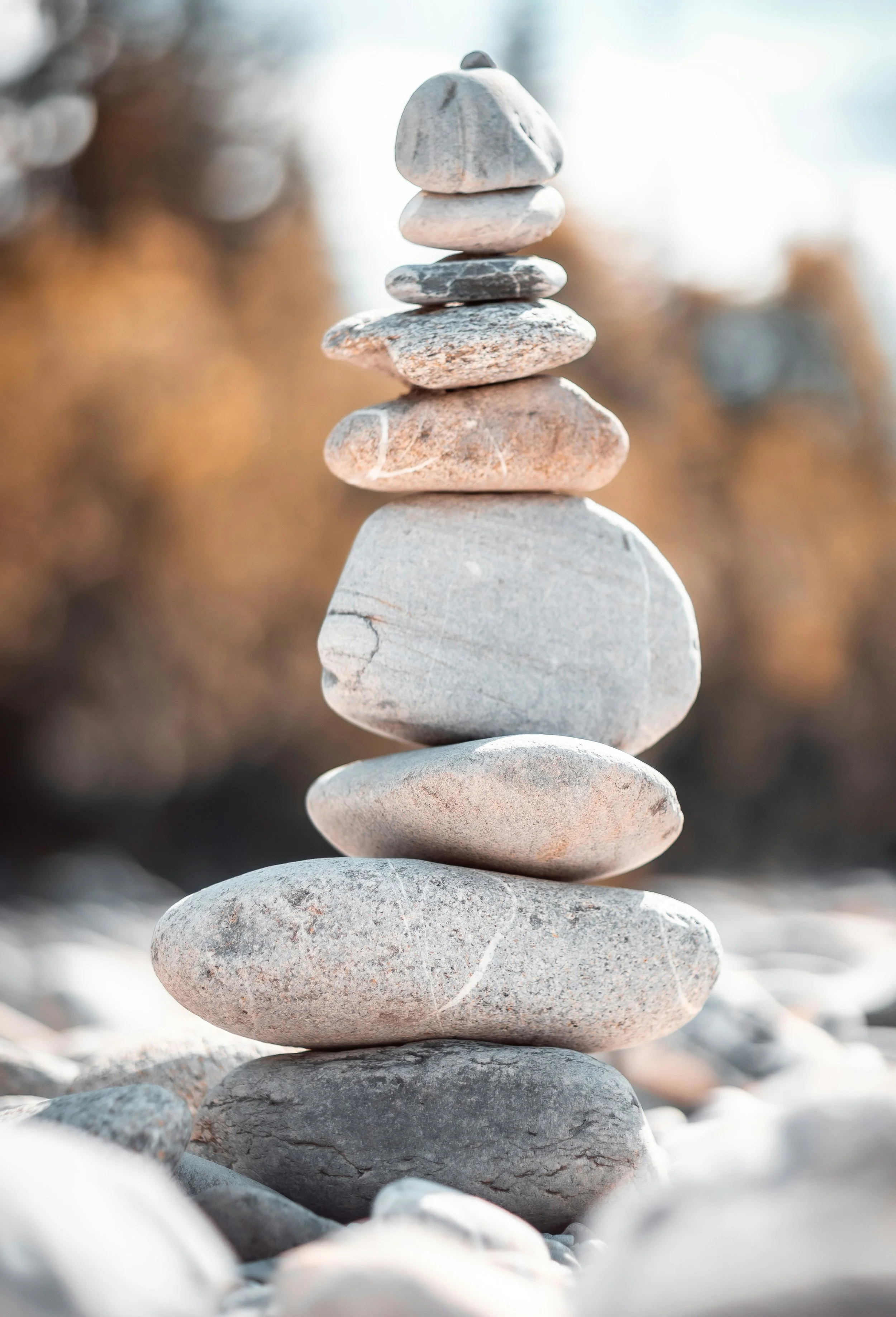 A stack of nine smooth, rounded stones of various sizes and colors, balanced on top of each other outdoors with sunlight in the background.