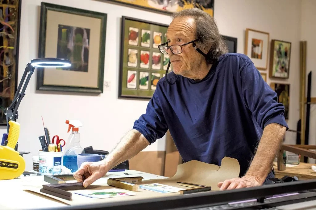 An elderly man with glasses and gray hair is working at an art studio, leaning over a table with framed art pieces and art supplies, including a desk lamp, spray bottle, and paintbrushes.