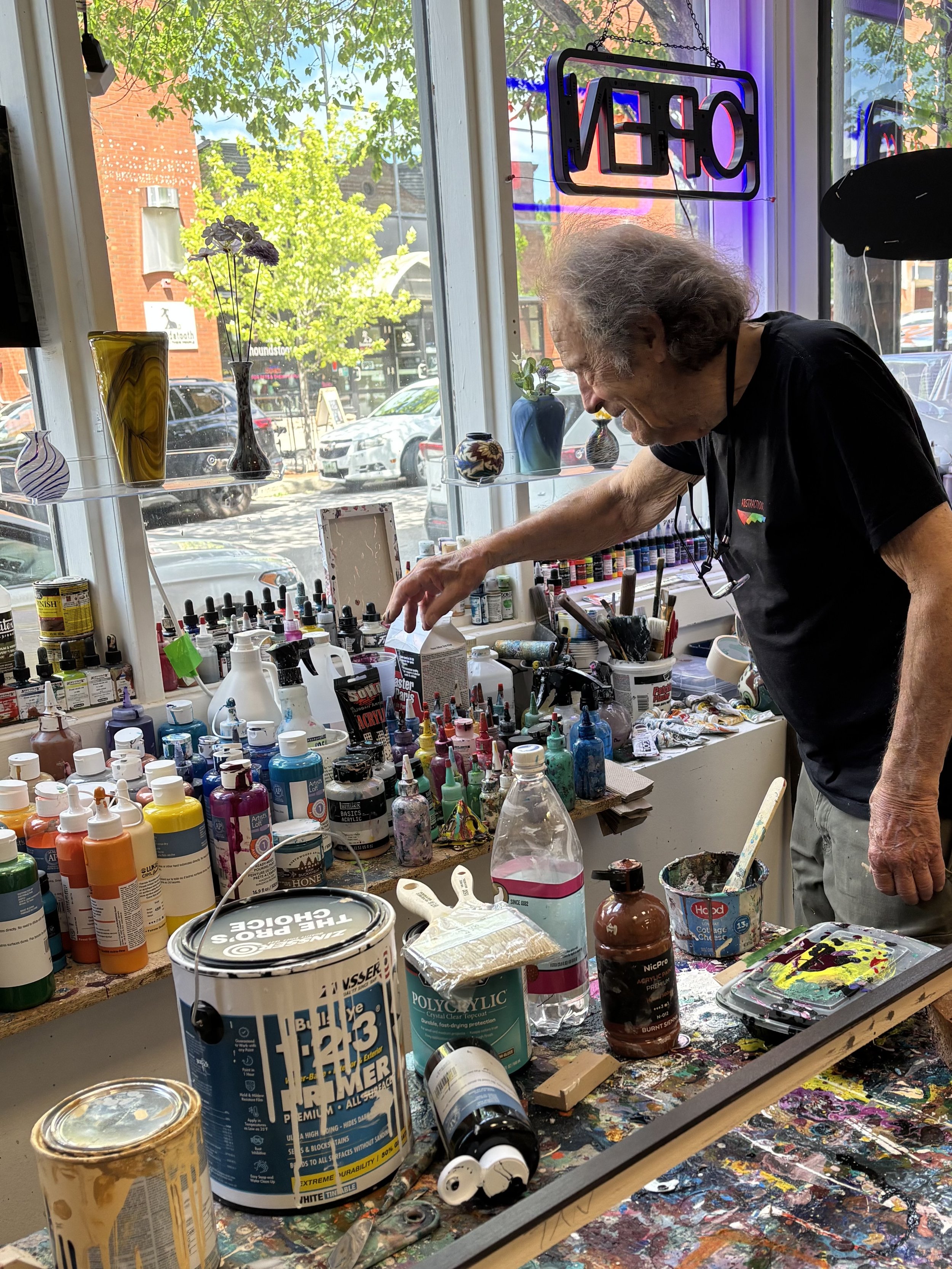 An artist's workspace filled with various art supplies, including numerous bottles of acrylic paint, brushes, and a palette with colorful paints. An elderly man is smiling and reaching for something, with a view of a sunny street and green trees outside the window.
