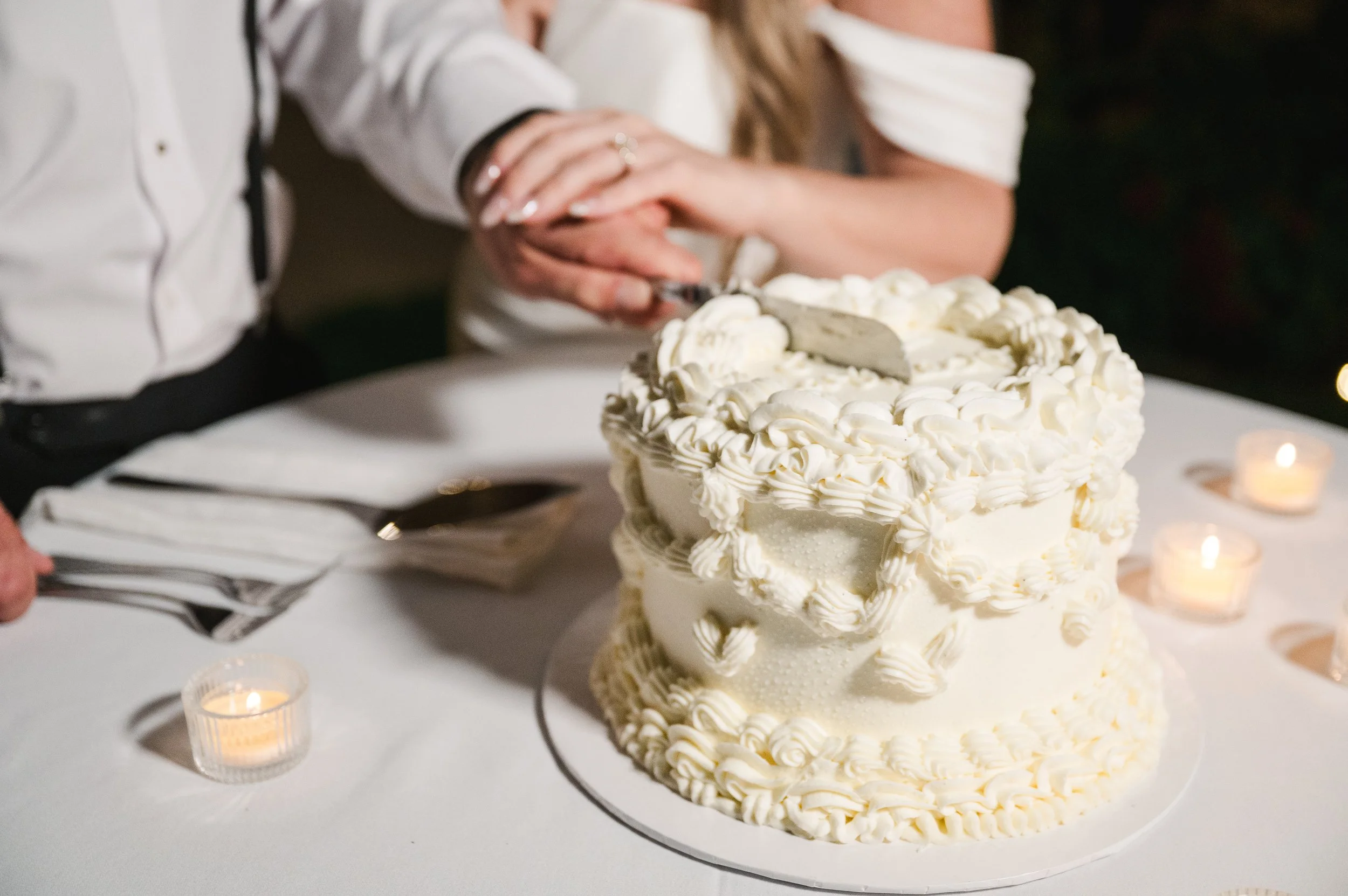 A person cutting a multi-layered wedding cake decorated with white frosting and piped icing accents.