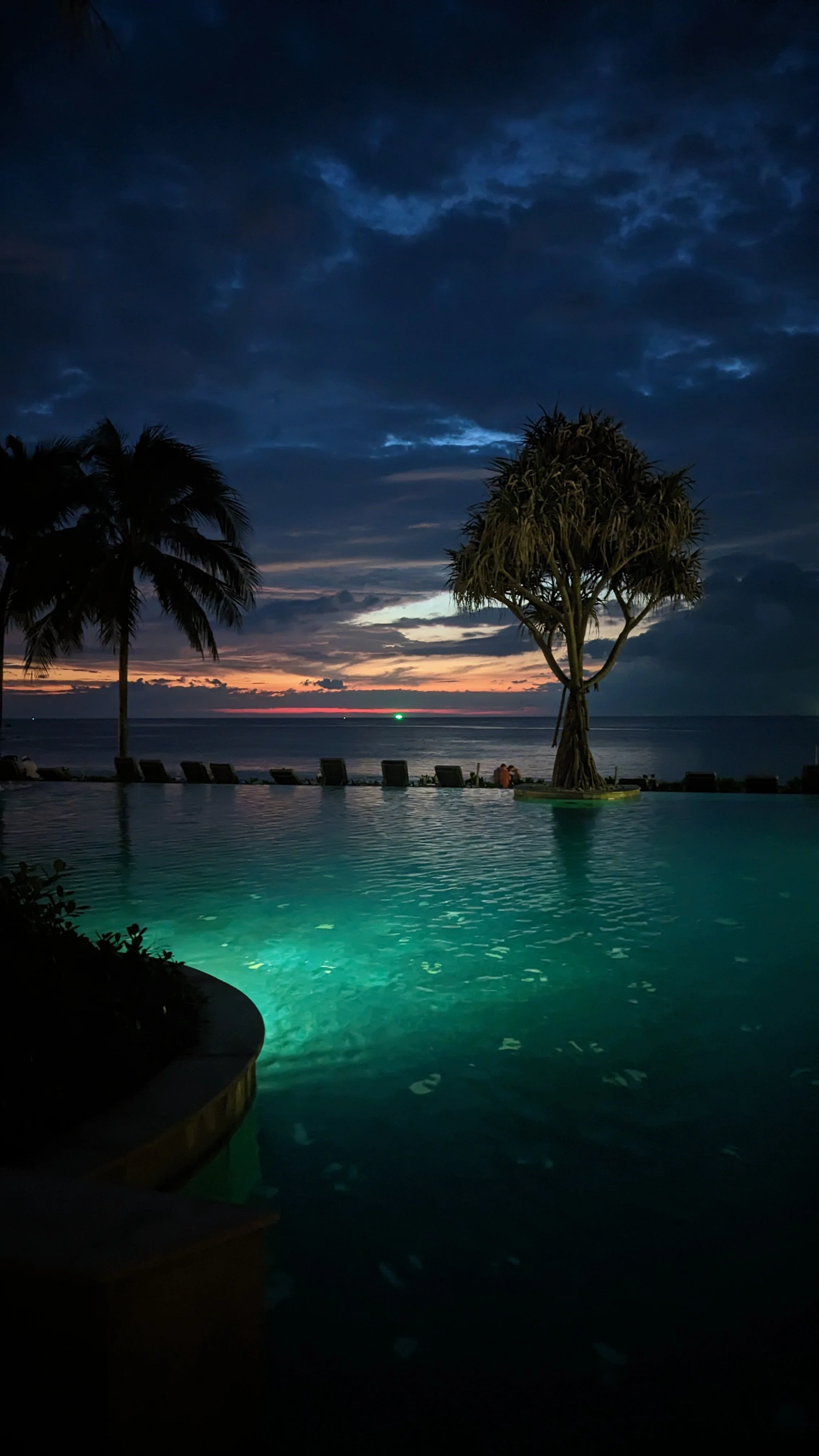 A nighttime view of a tropical scene featuring a pool illuminated with underwater lights, two palm trees, a single tree, lounge chairs along the beach, and a colorful sunset sky with dark clouds over the ocean.