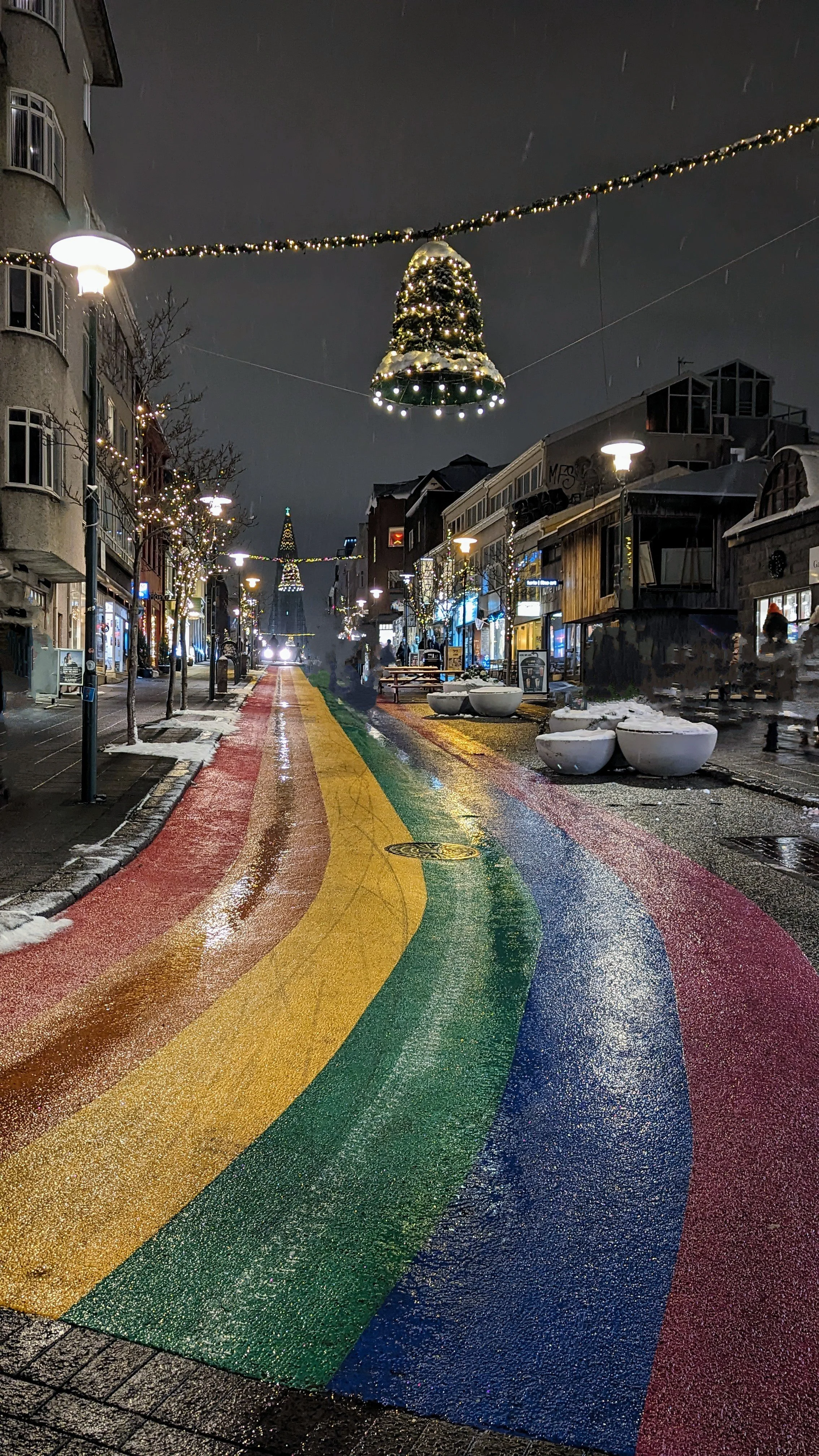 A city street decorated for Christmas with a rainbow-colored crosswalk, Christmas lights, and a lit Christmas tree overhead during a rainy night.
