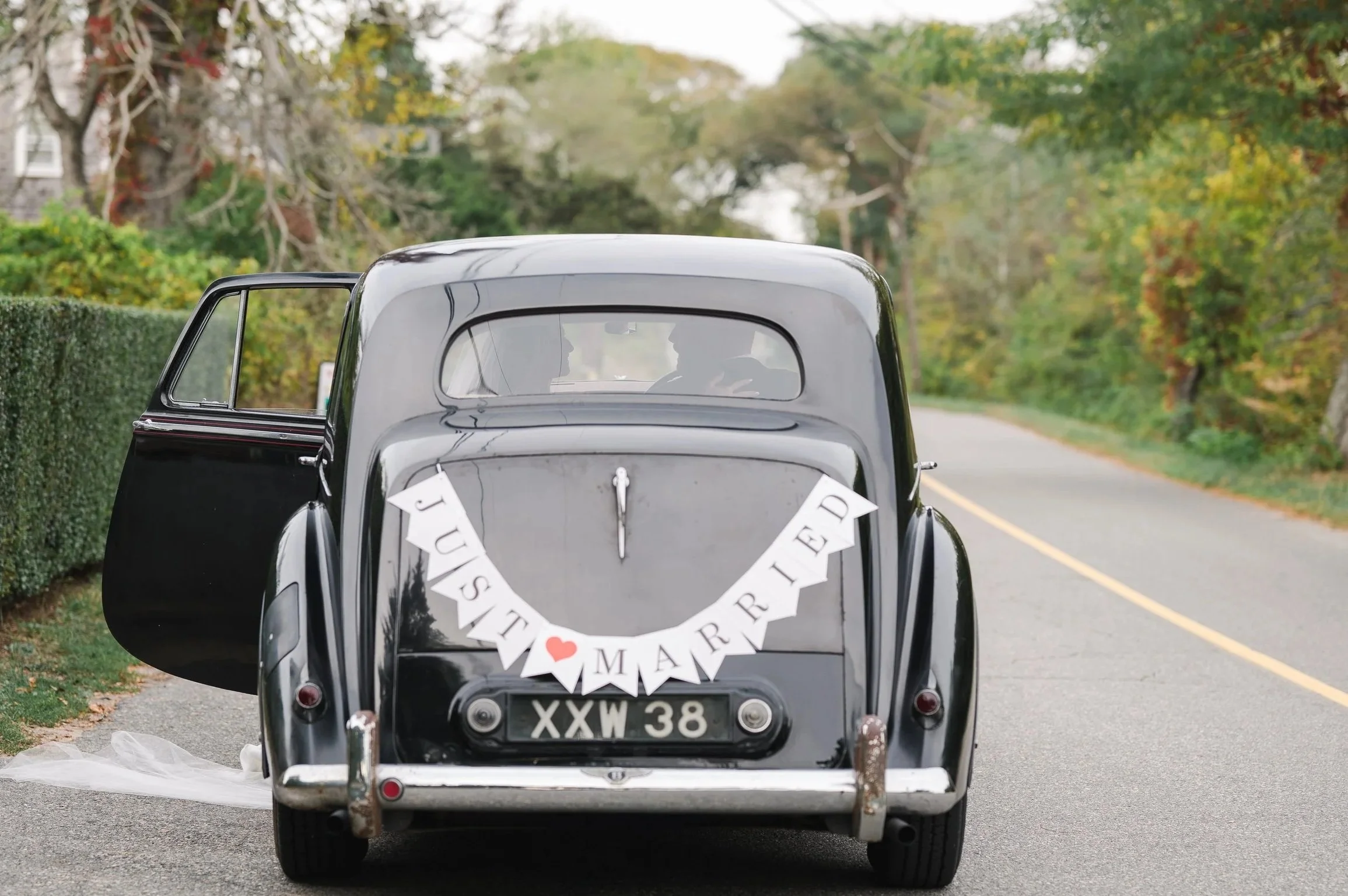 A vintage black car with its door open on a rural road, decorated with a 'Just Married' banner on the rear window, surrounded by trees with autumn foliage.