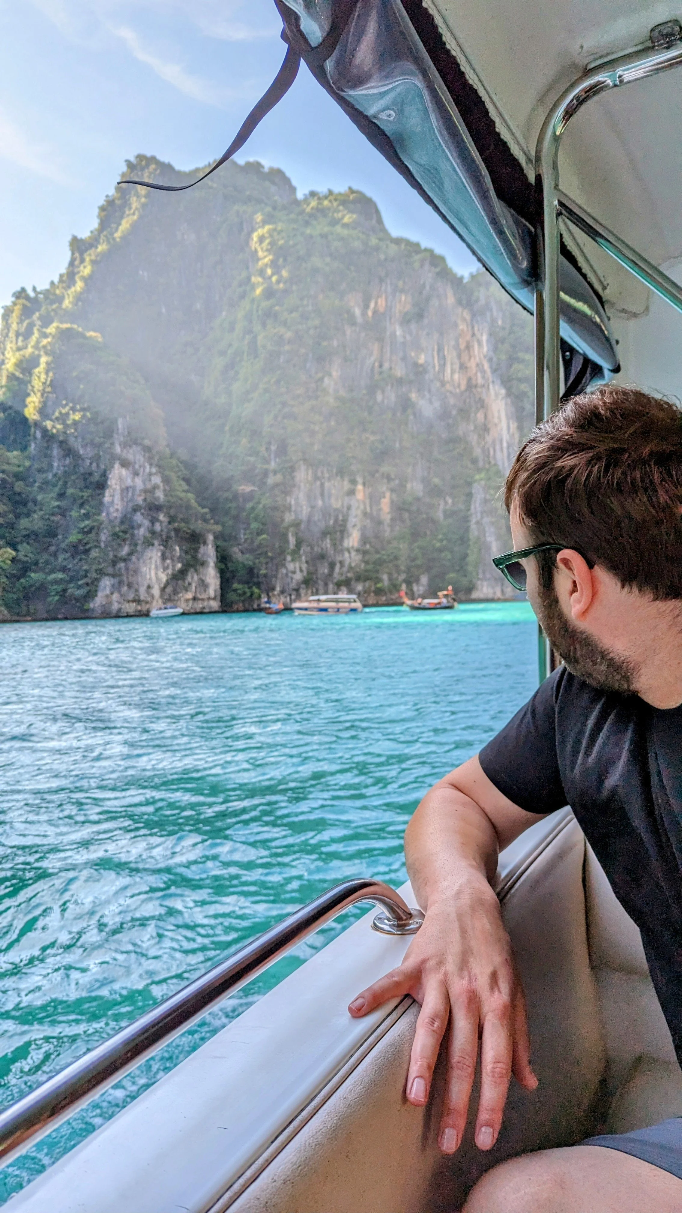 A man with sunglasses leaning on the side of a boat, looking at a scenic view of cliffs and green islands in the distance over blue water.