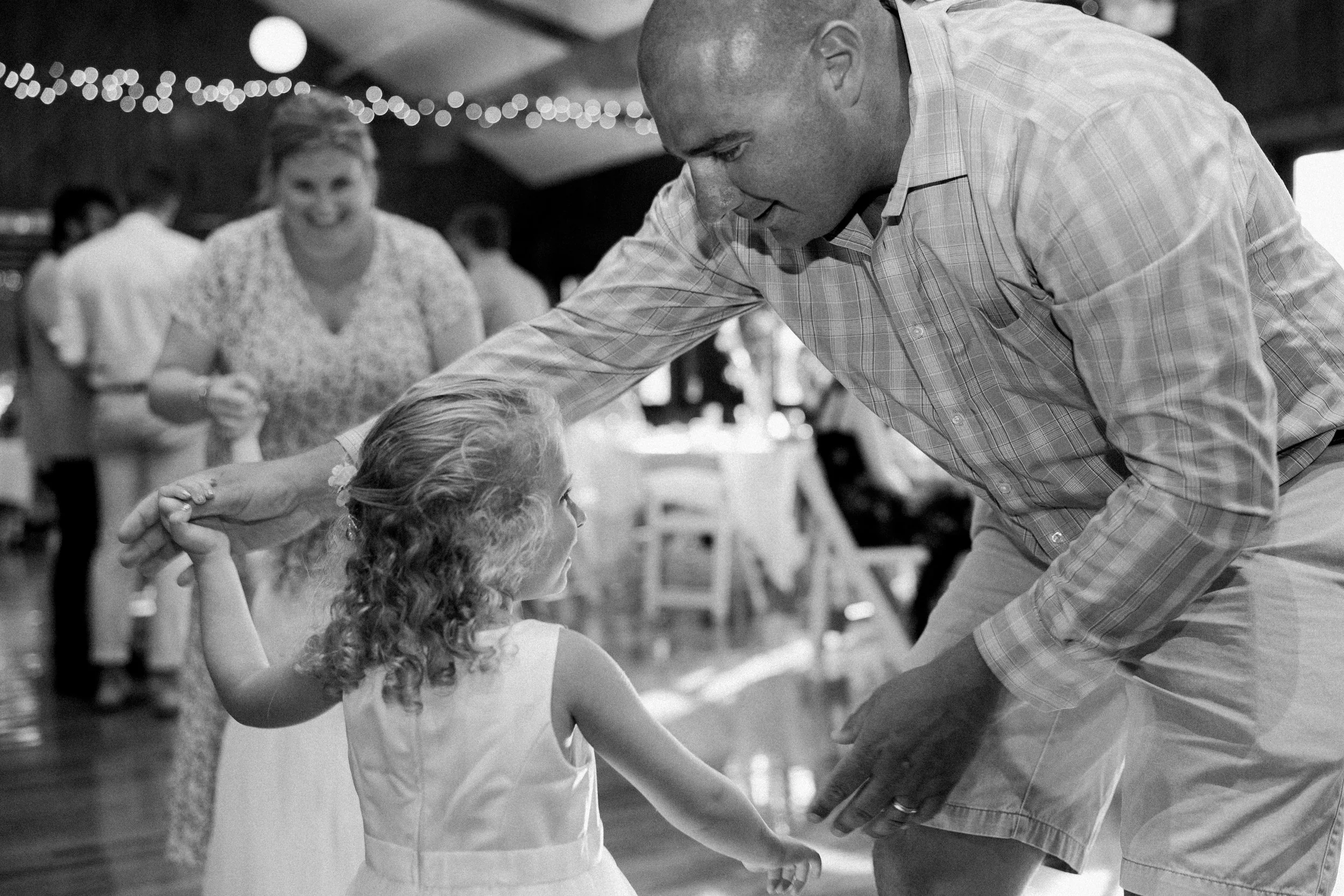 A man is dancing with a young girl at a social event, holding her hands. In the background, there are other people, including a woman smiling and observing the dance, with string lights hanging overhead.