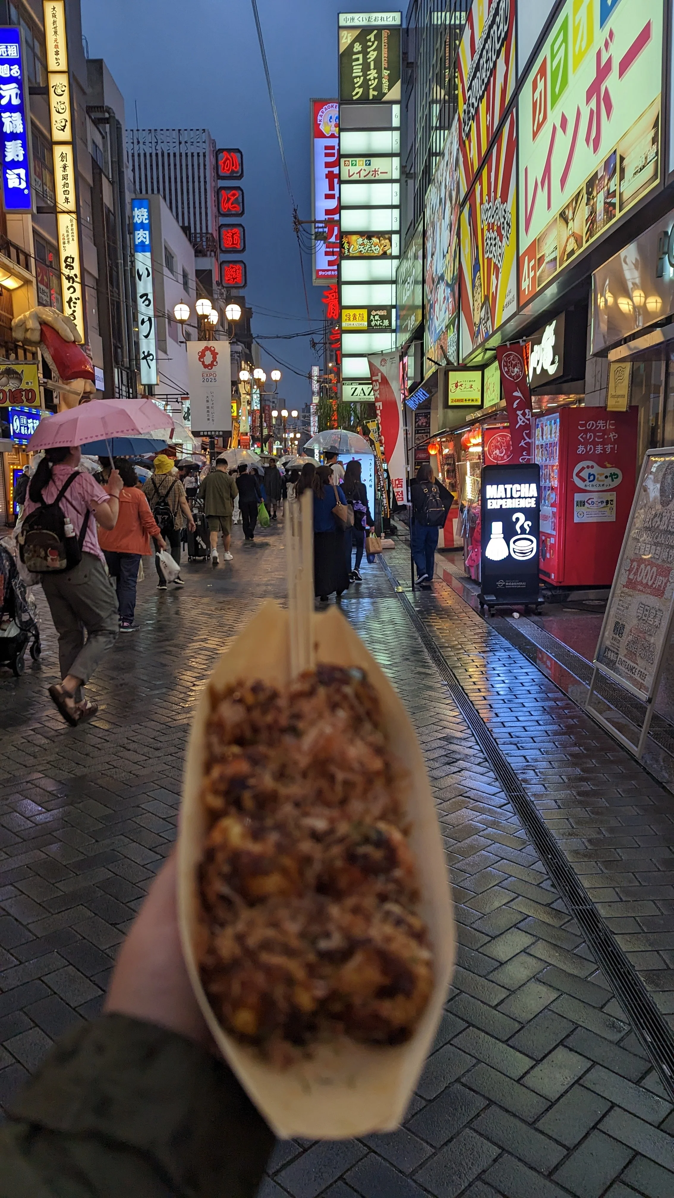 A street scene in Japan during the evening with many people walking, illuminated signs, and buildings with colorful advertisements. The person in the foreground is holding a snack on a stick, with the focus on the snack and the background showing lively city life.