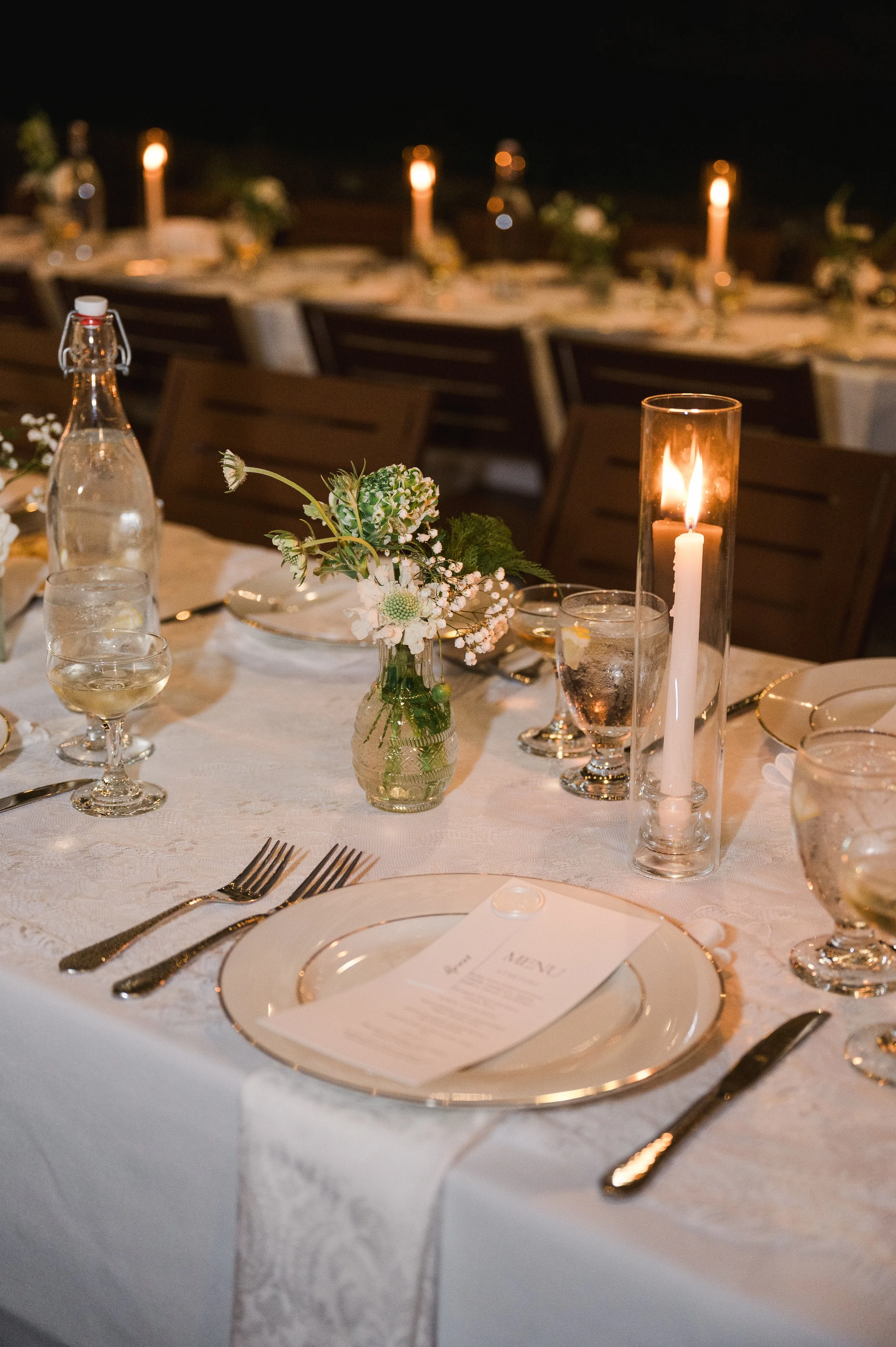 Elegant dining table set for a special occasion, illuminated by candlelight with floral arrangements and glassware, in a dimly lit setting.