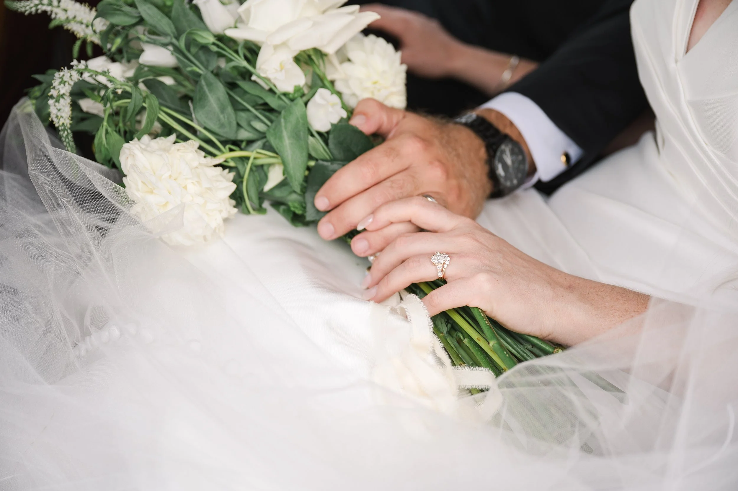 Close-up of a newlywed couple's hands, with wedding rings, holding a bouquet of white flowers and greenery.