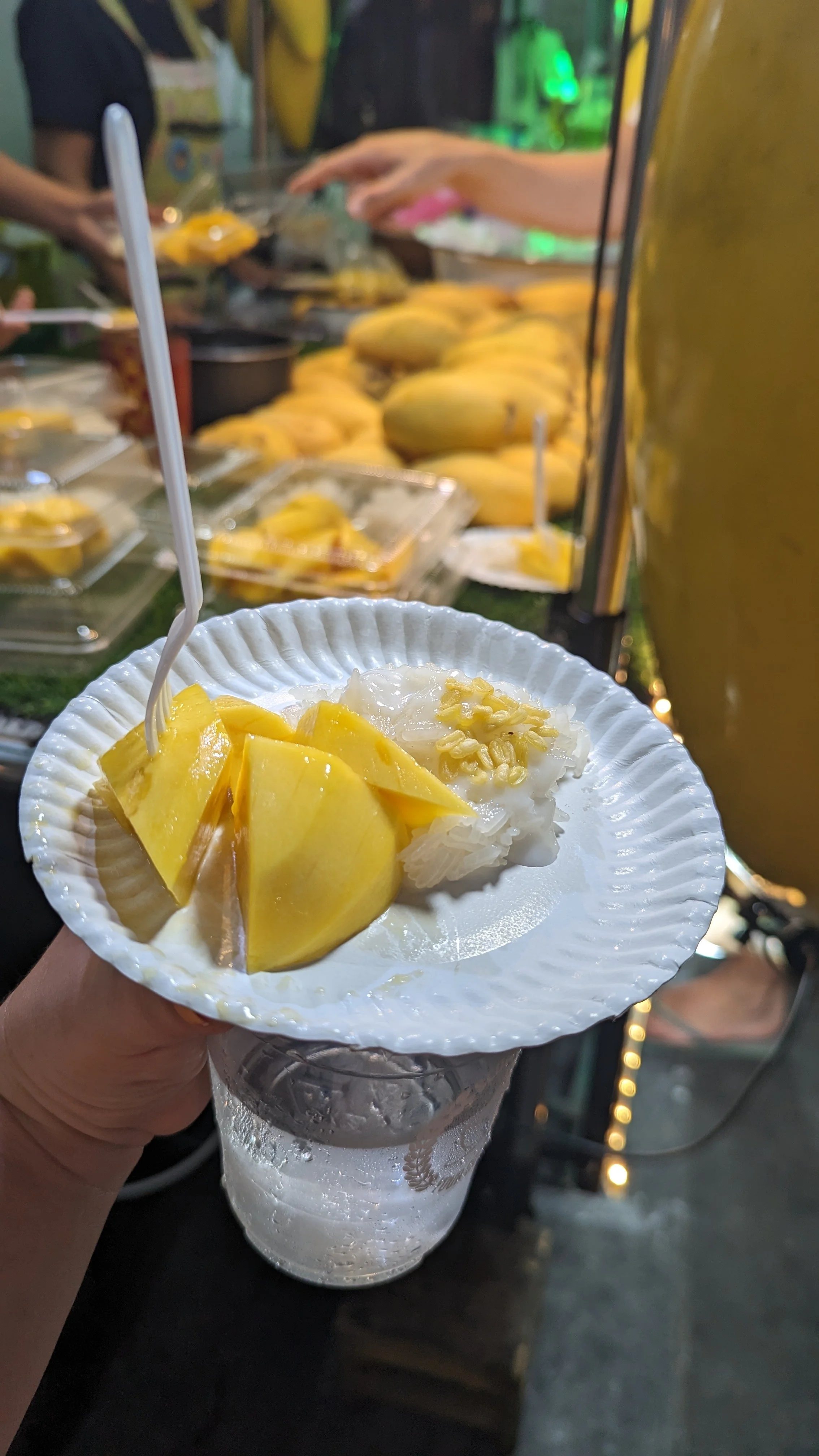 Close-up of a dessert with yellow mango slices and white sticky rice, served on a clear glass dish with a raised pattern. The background shows an assortment of mangoes and other food items at a market stall.