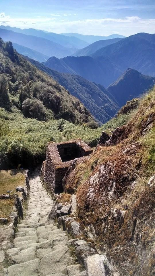 Stone steps leading up a mountain trail, with a small stone structure on the side, overlooking a lush green valley and mountain ranges in the distance.