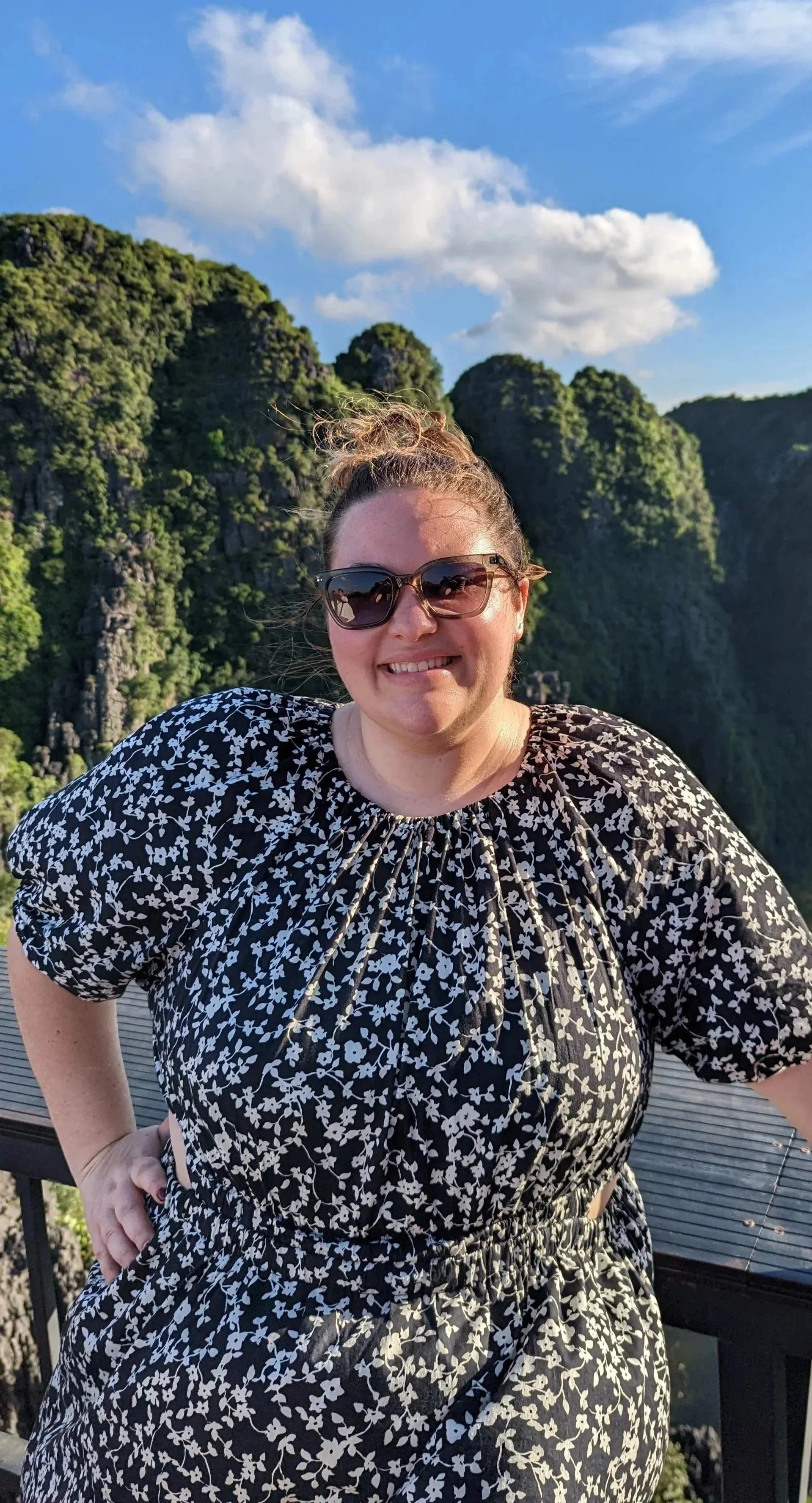 A woman with curly hair in a bun wearing sunglasses and a black and white floral dress with puffed sleeves, standing outdoors with lush green mountains in the background and a partly cloudy sky.