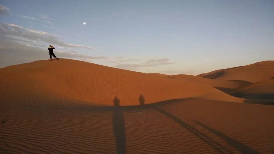 A person standing on a sand dune and taking a photo in a desert at sunset, with long shadows and a visible moon in the sky.