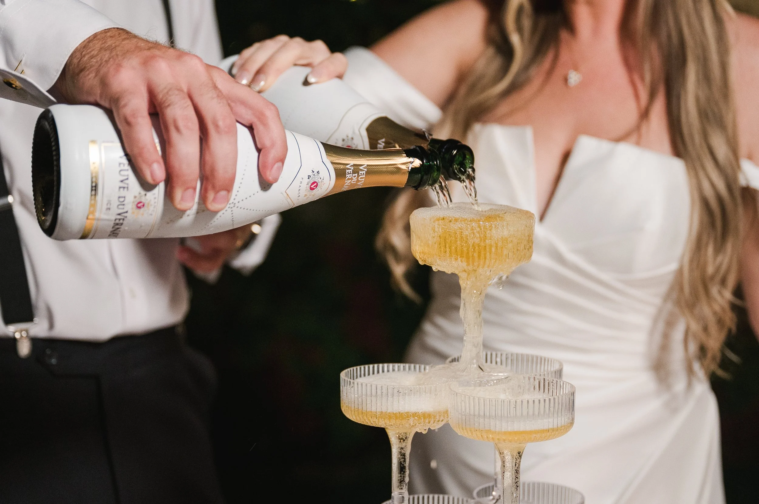 A person in a white shirt pours sparkling wine from a bottle into a glass, which is placed on a tiered stand with other glasses beneath.