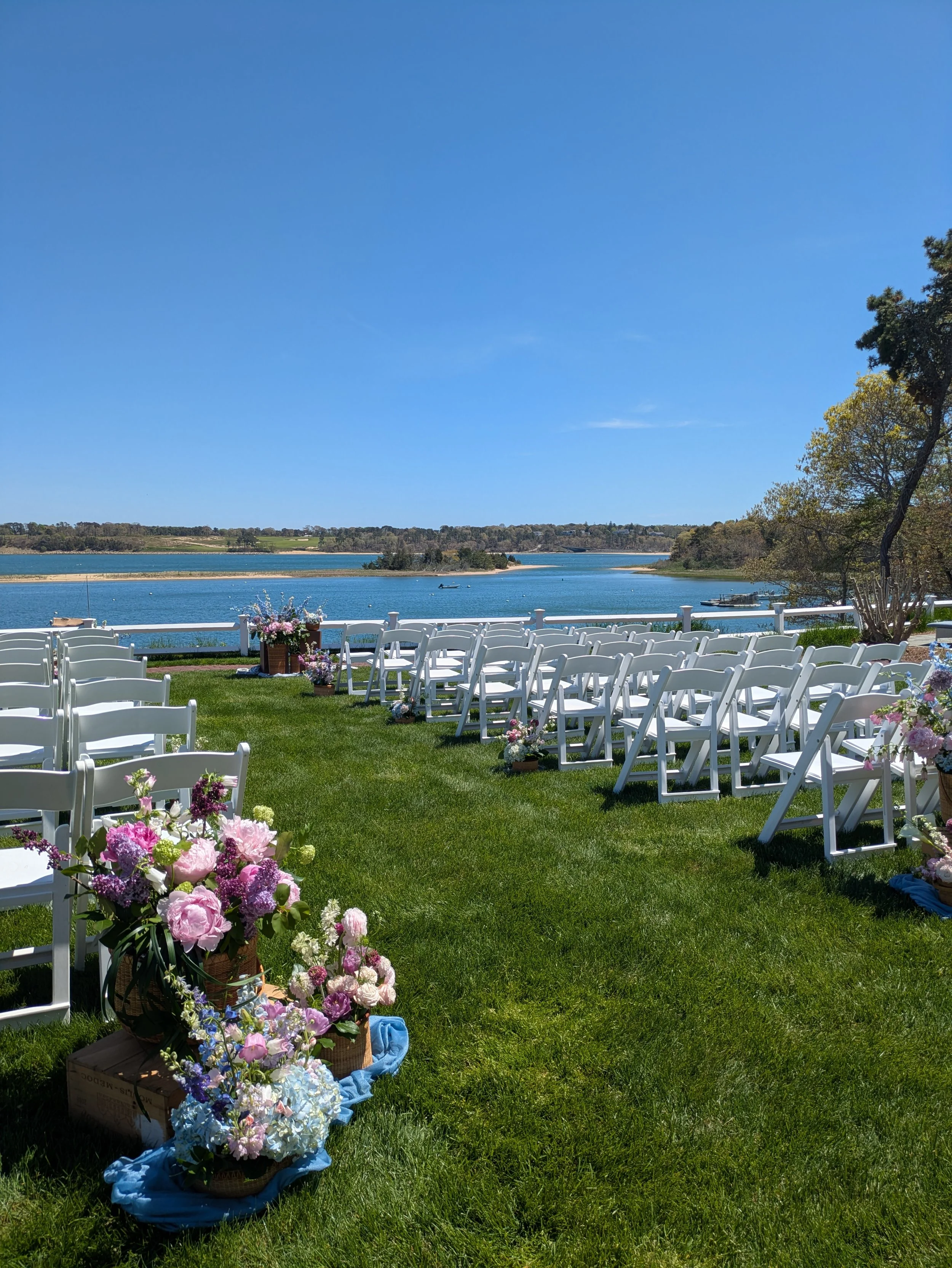 Outdoor wedding setup with white chairs arranged on a grassy area facing a scenic view of a river or lake, with pink, purple, and white flowers in baskets and potted plants in the foreground, trees on the right, and a clear blue sky overhead.