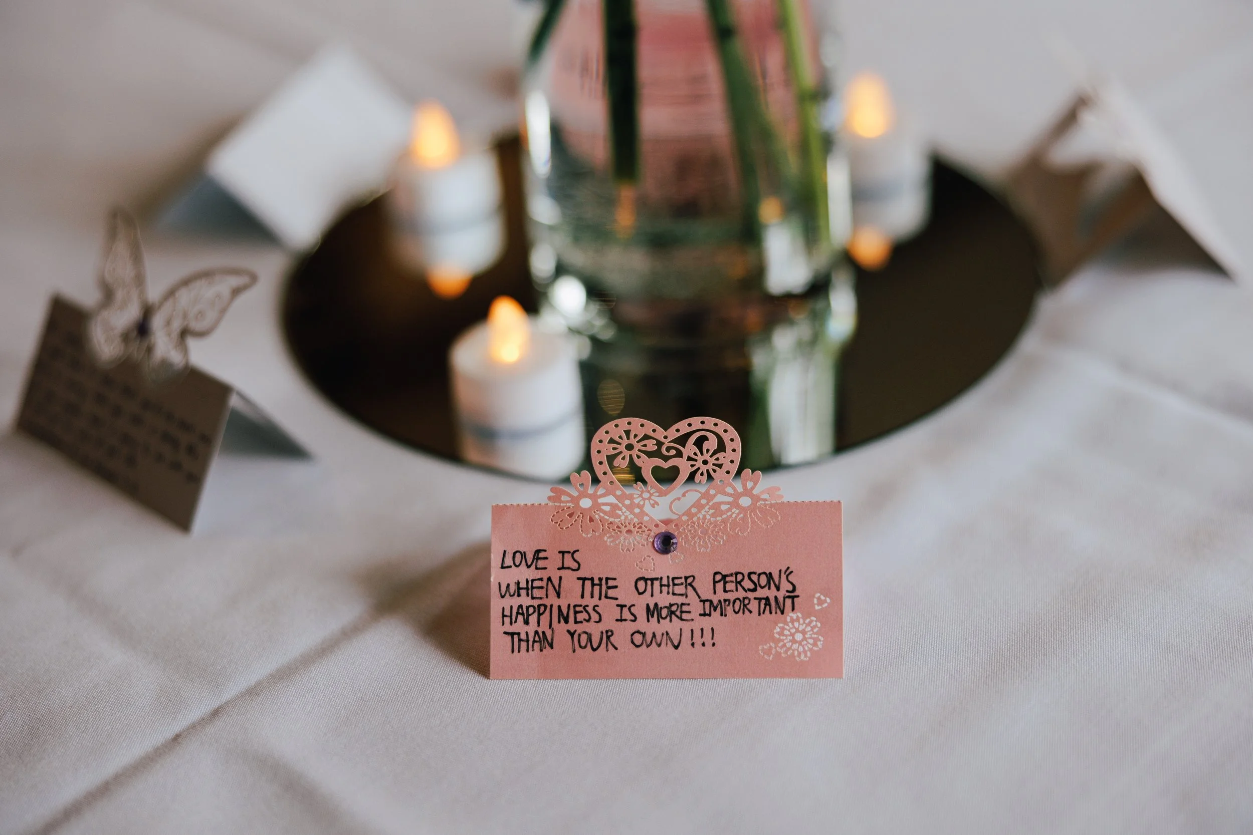 A pink card with a decorative floral cutout at the top, with the handwritten quote: "Love is when the other person's happiness is more important than your own!!!". The card is on a white table surrounded by small lit candles and decorative paper butterflies.