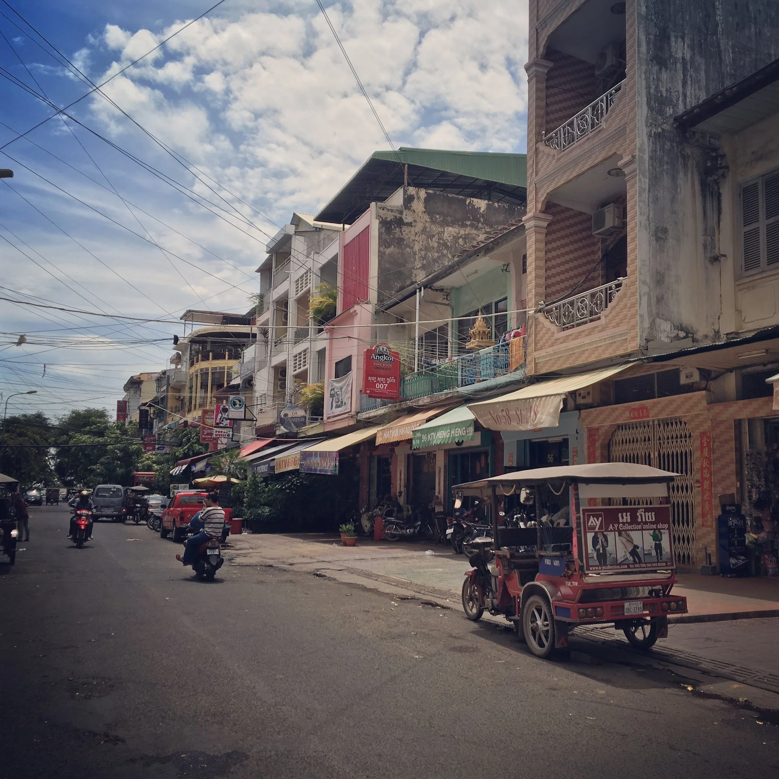 Street scene with multistory buildings, parked motorcycles and cars, people riding motorcycles, shops with signs, and overhead power lines under a cloudy sky.