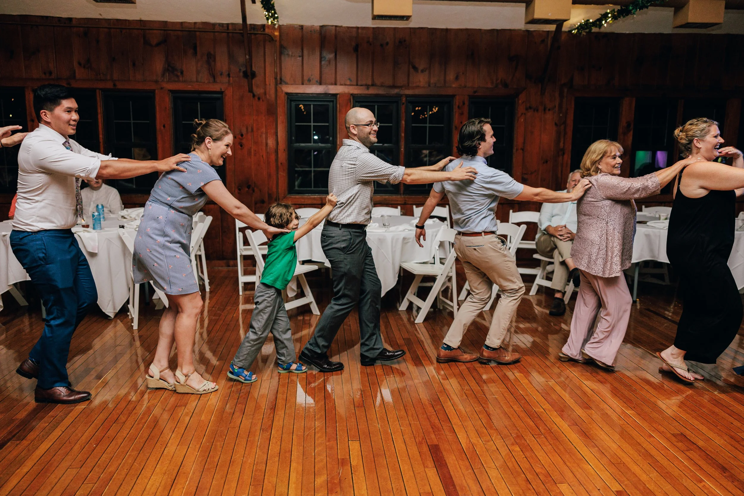 People at a celebration or party engaging in a conga line dance indoors, with wooden walls and string lights in the background.