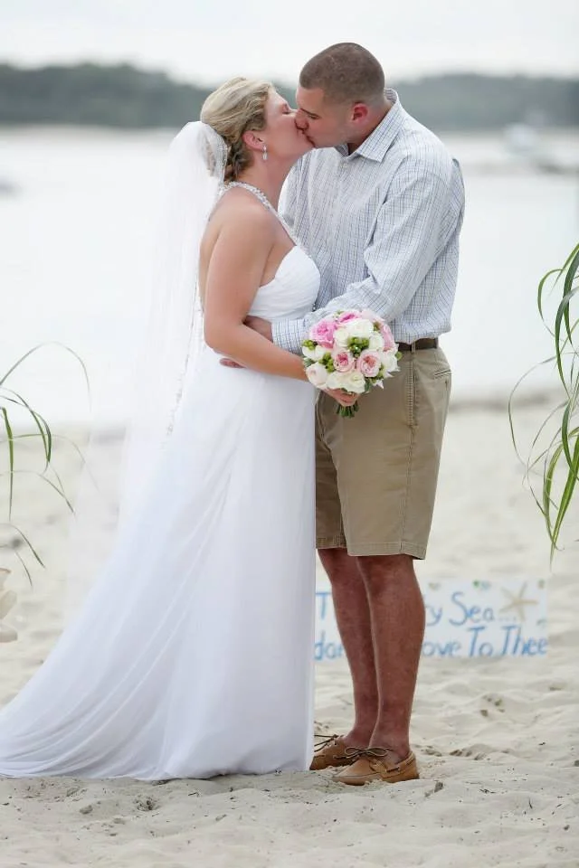 A bride and groom kissing during a beach wedding, with the bride holding a pink and white bouquet and a backdrop of water and sand.