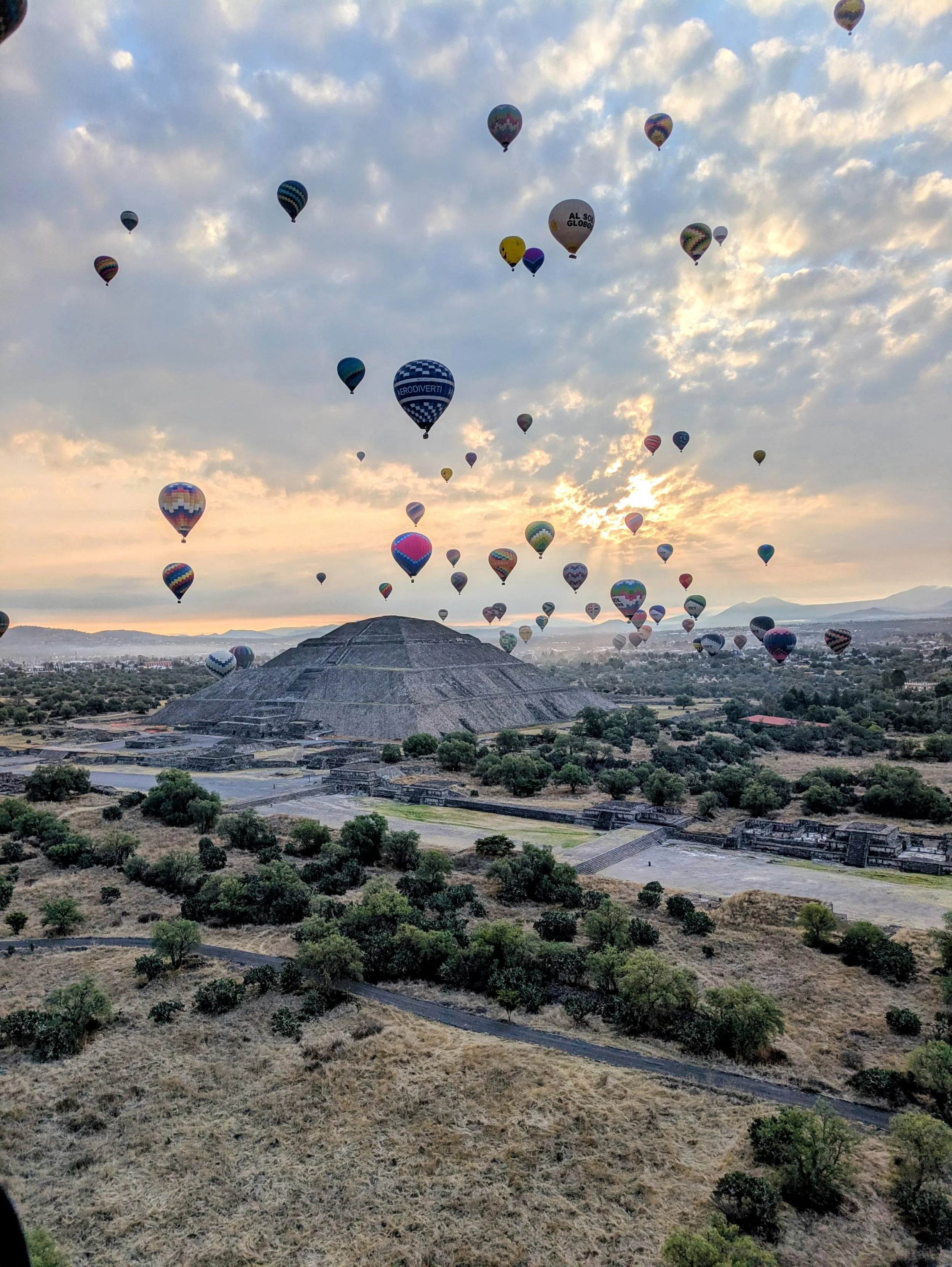 Multiple hot air balloons floating over ancient Mayan ruins and a landscape at sunrise or sunset.