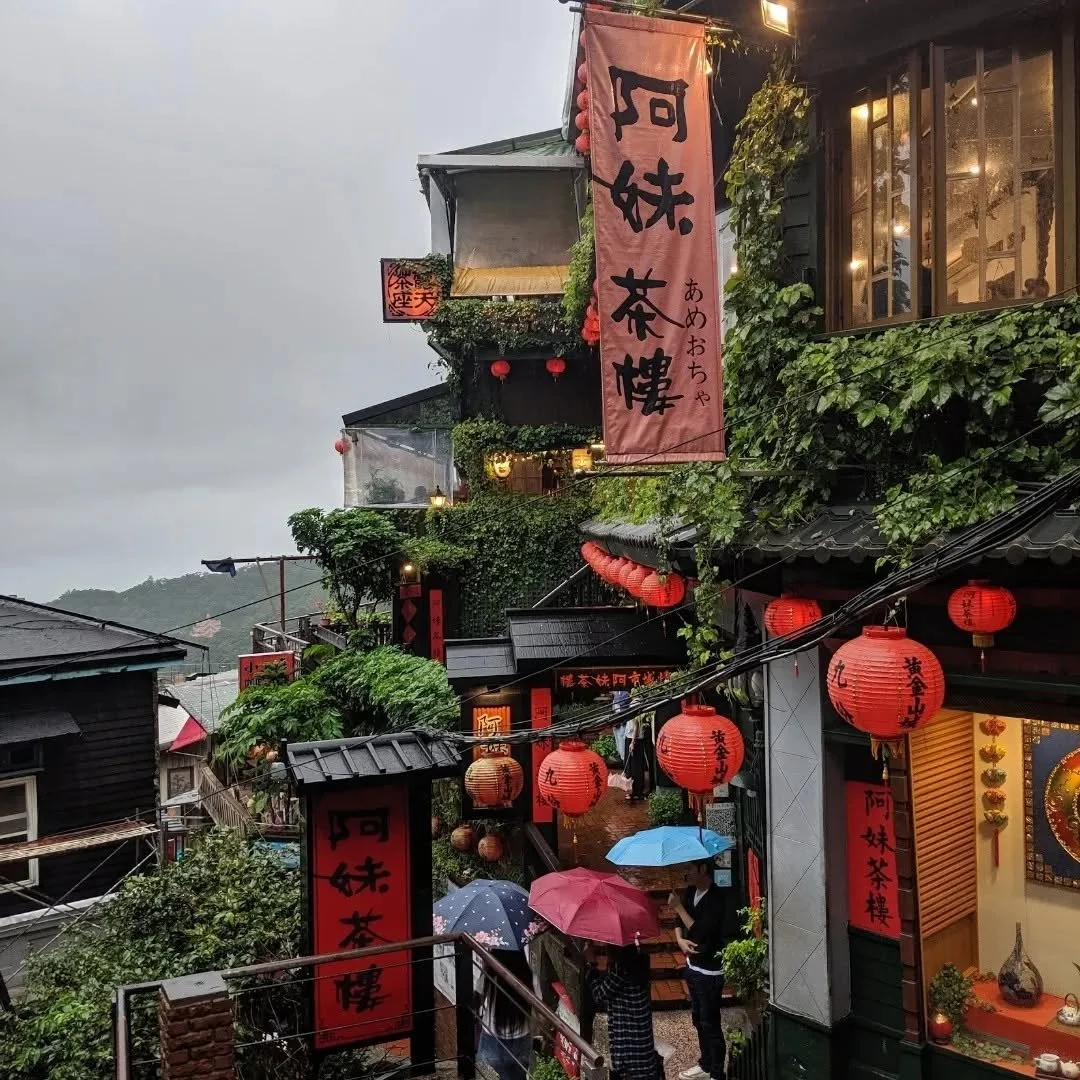 Street view of a traditional Asian-style building decorated with red lanterns, illuminated signs, and greenery, with people holding umbrellas.