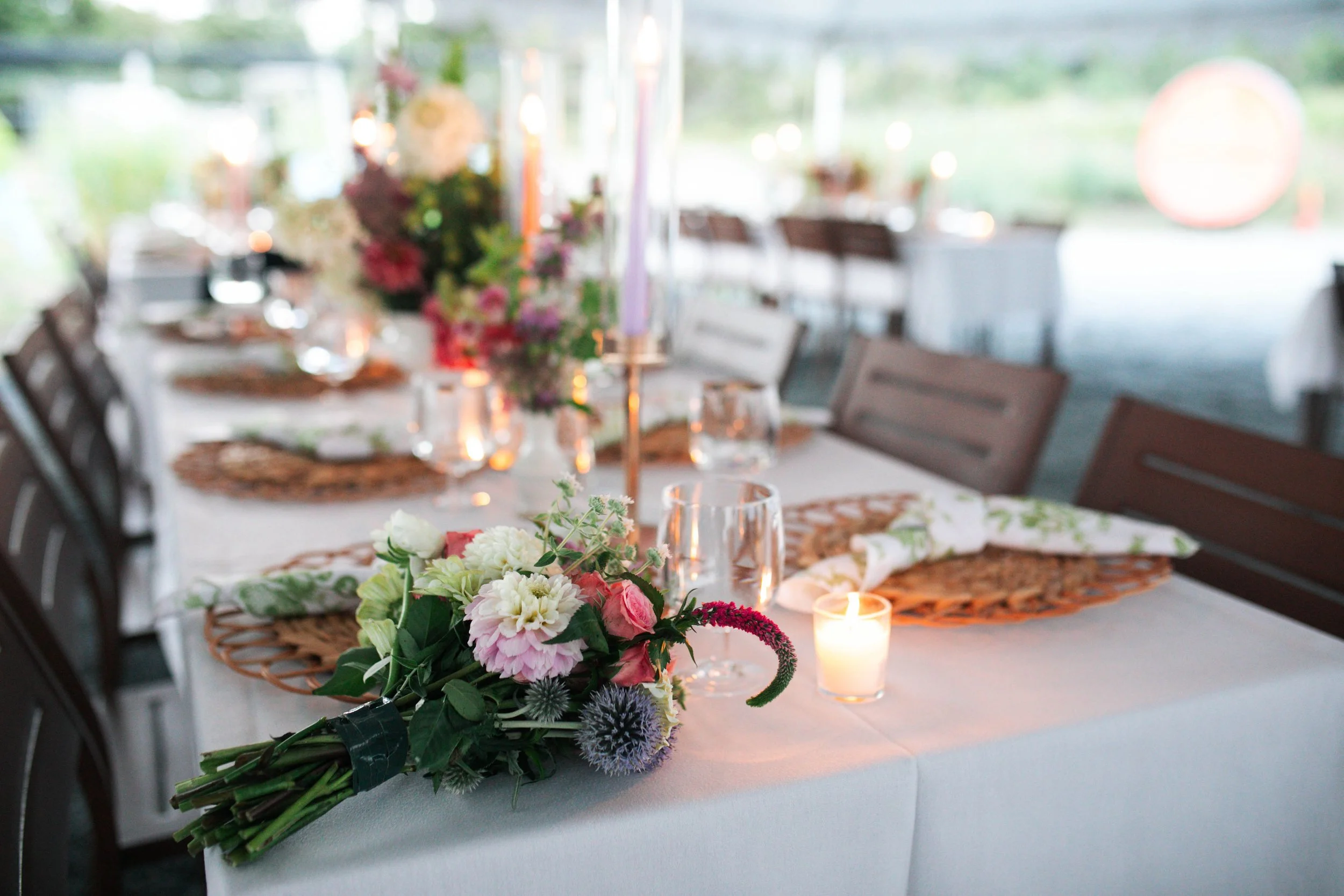 A table decorated with a bouquet of pink, white, and purple flowers, surrounded by glasses and lit candles, set in a bright, airy room with large windows and greenery outside.