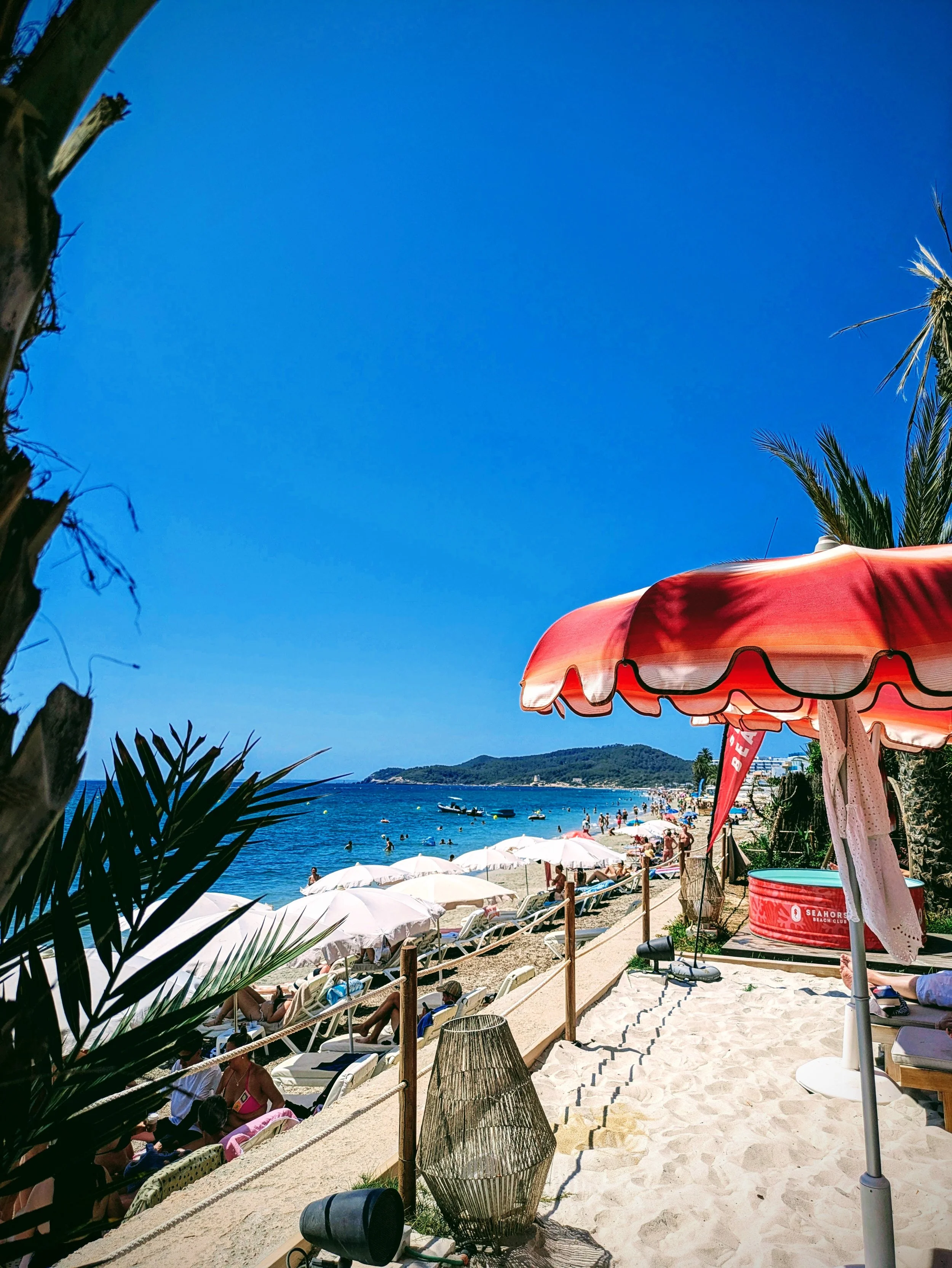 A crowded beach with umbrellas, lounge chairs, and people swimming in the ocean, viewed from under a red and white beach umbrella with palm trees and hills in the background.