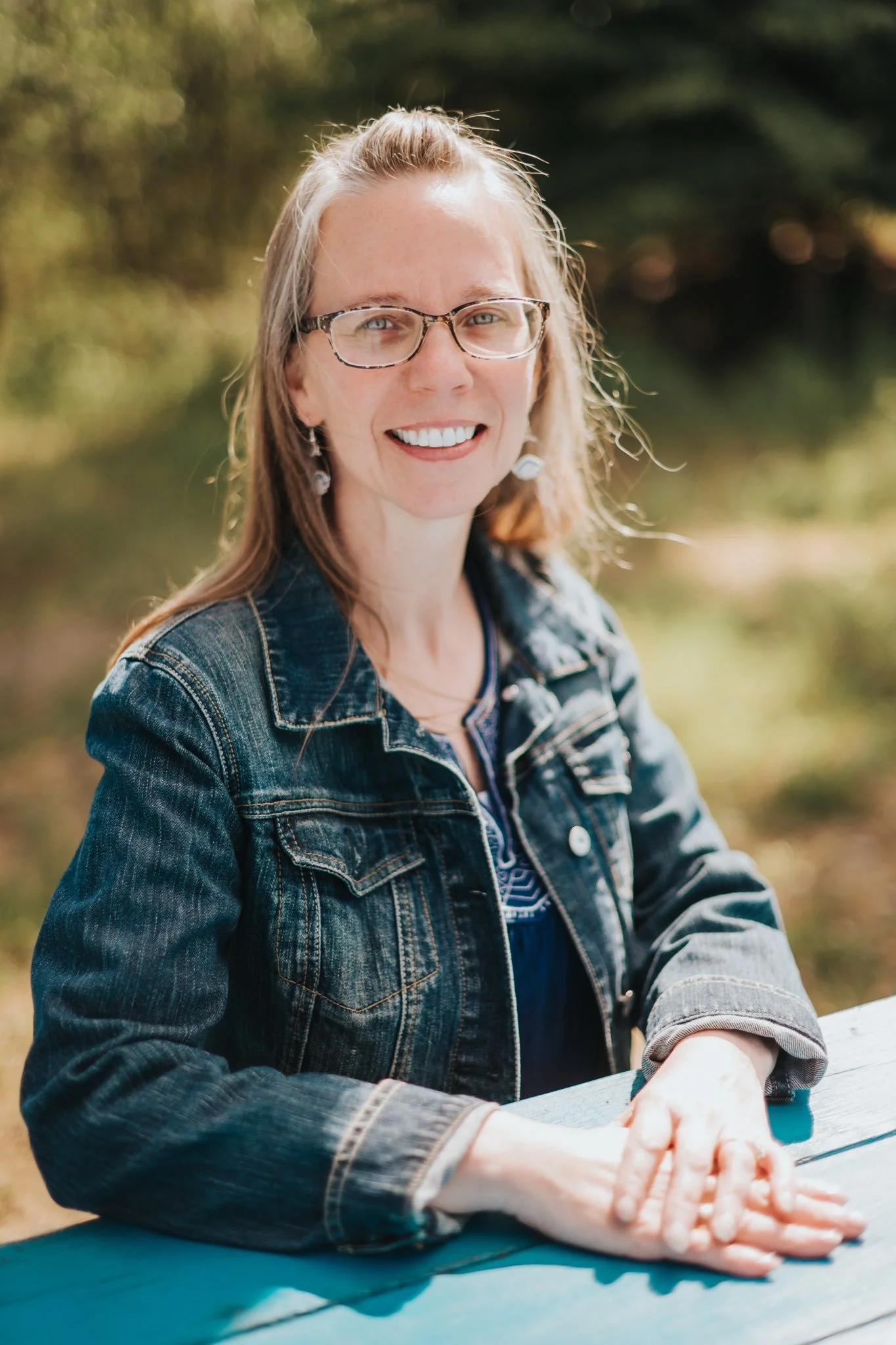 A woman with glasses and earrings, wearing a denim jacket, smiles while sitting outdoors in a green area.