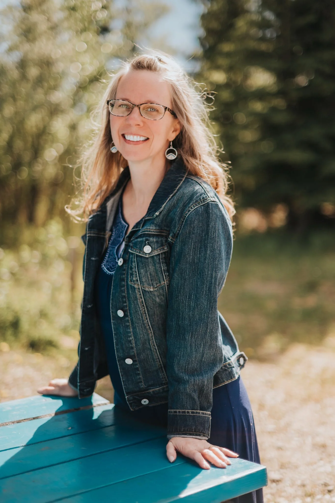 A woman with long blonde hair, wearing glasses and a denim jacket, smiling outdoors with blurred trees in the background.