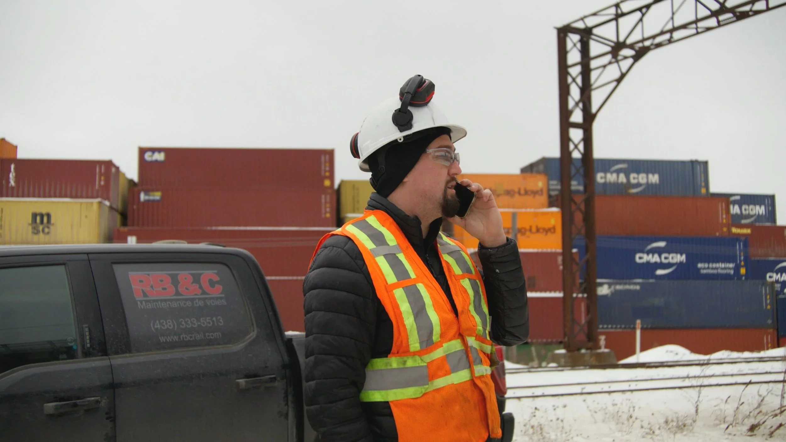 Un homme portant un casque et un gilet de sécurité orange parle au téléphone dans une zone de stockage avec des conteneurs dans un port.