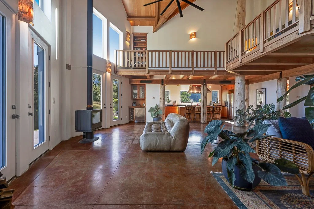 Interior view of a spacious living room with high ceilings, wooden beams, and large windows letting in natural light. There is a leather sofa, indoor plants, and a view of the kitchen and dining area in the background.