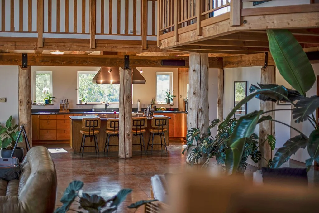 Open-concept kitchen with wooden cabinetry, island, bar stools, and large windows, in a home with exposed supporting beams and wood accents.