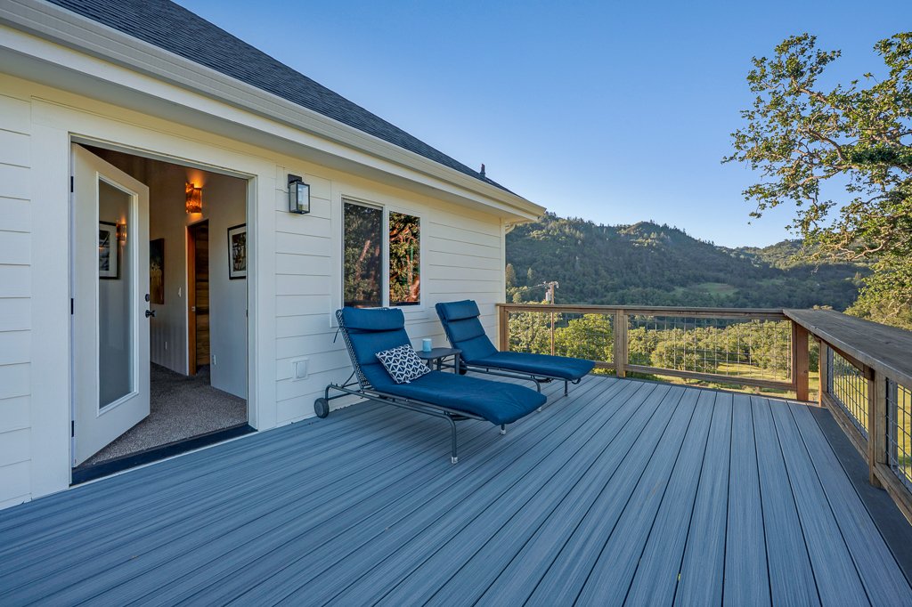 Two blue lounge chairs with a small pillow on a large wooden deck outside a white house, overlooking a scenic landscape with trees and mountains.