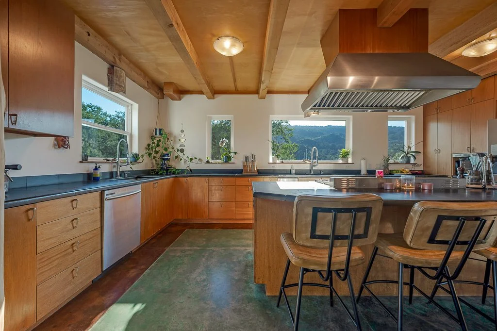 A kitchen with wooden cabinets, a central island with barstools, large windows showing a scenic outdoor view, and a modern range hood above the cooktop.