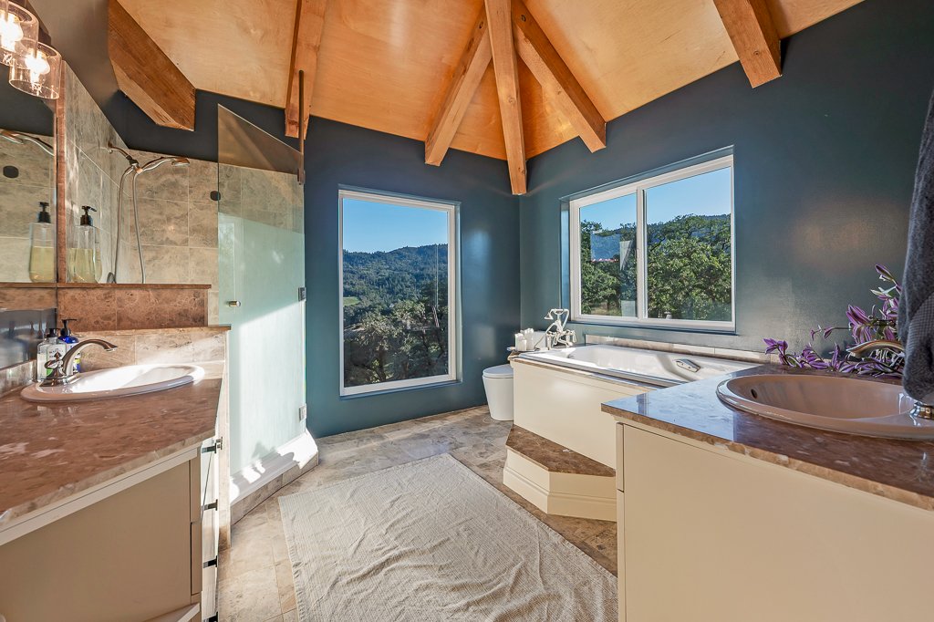 Bathroom with a skylight ceiling, two windows with scenic mountain view, corner bathtub, double sinks with marble countertops, and beige tile flooring.