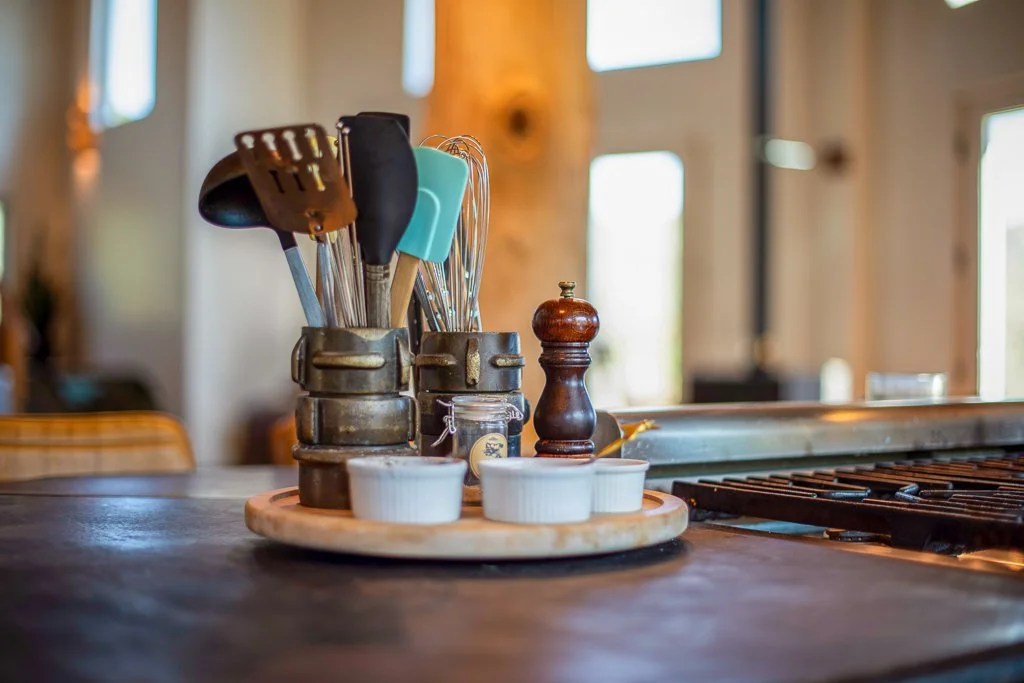A wooden tray with white ramekins, condiments, and kitchen utensils on a kitchen counter.