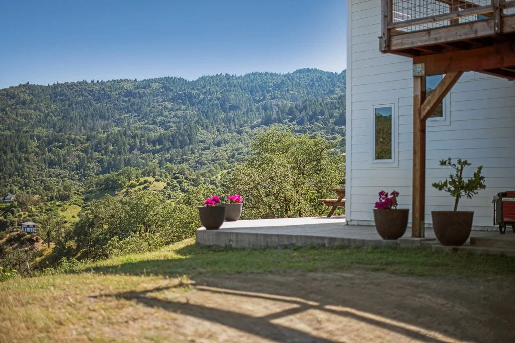 Part of a white house with a small balcony or deck, potted plants with pink flowers, a picnic table, and a view of rolling green hills and trees under a blue sky.