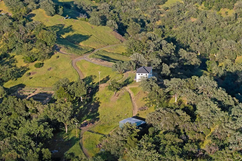 Aerial view of a rural landscape with a white house, unpaved roads, green fields, wooded areas, and a small pond surrounded by trees.