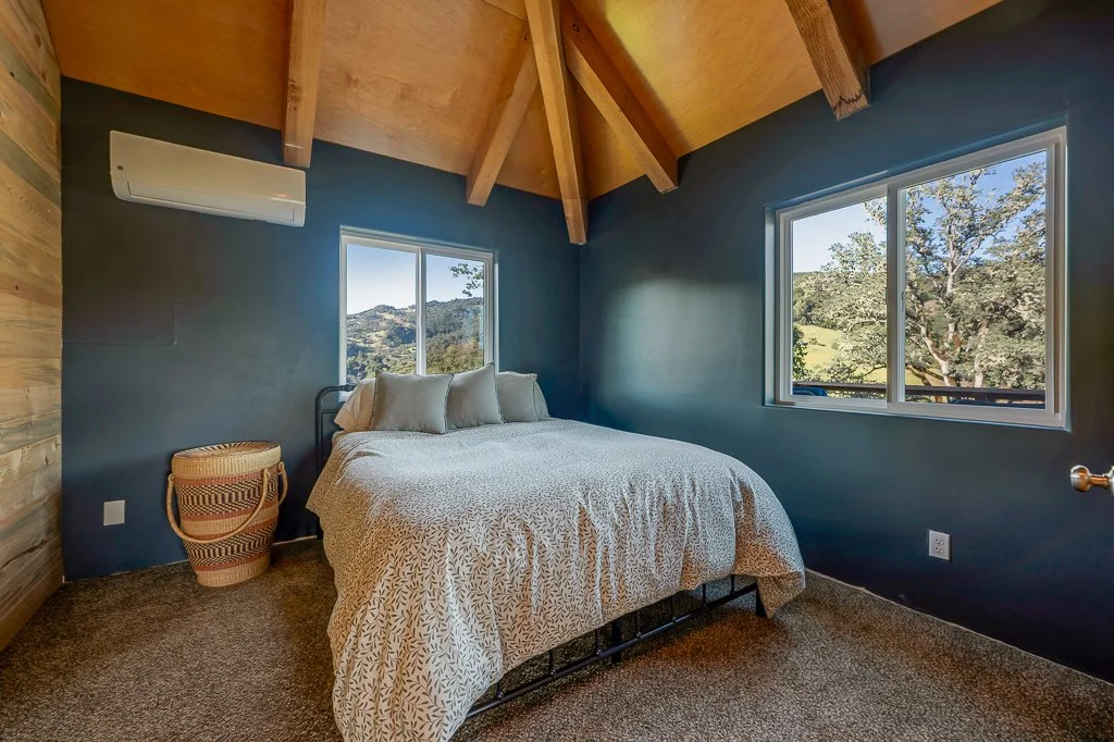A cozy bedroom with a wooden ceiling, a window showing a scenic view of trees and hills, a bed with neutral bedding, an air conditioning unit, and a basket near the bed.