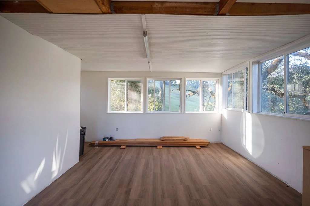 Empty room with large windows, wood flooring, white walls, and a ceiling with exposed wood beams, with construction materials on the floor.
